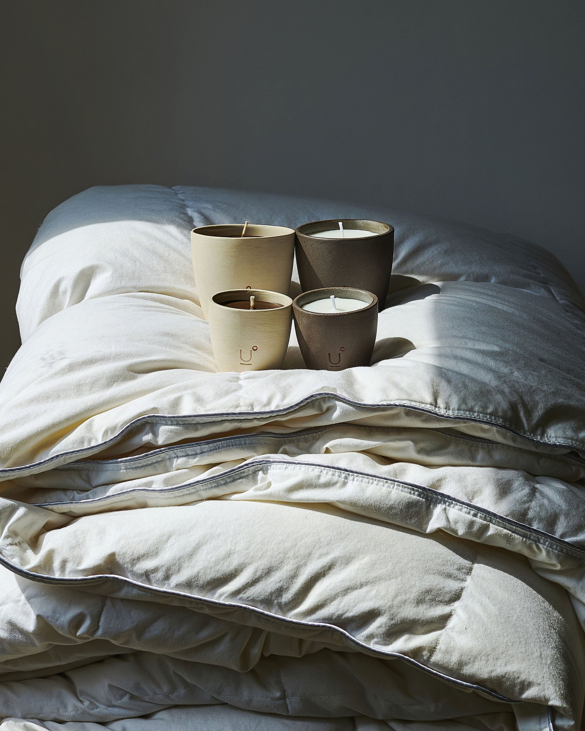 Four candles in minimalist ceramic holders, placed on a bed with white bedding and a pillowy comforter. YOUTH OF LIGHT HANDMADE CANDLES