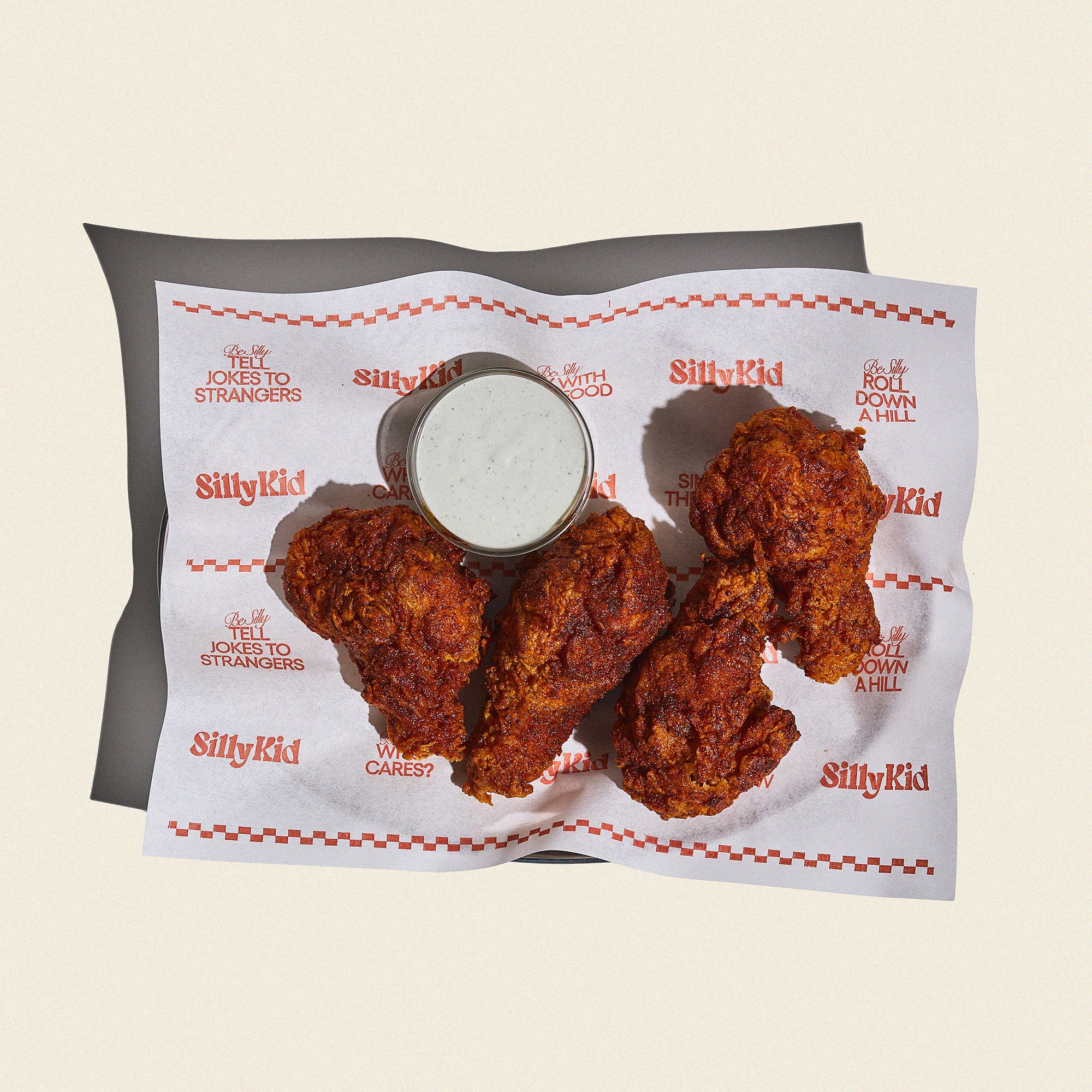 Three pieces of fried chicken with a side of ranch dipping sauce on branded paper with red text and checkered pattern.