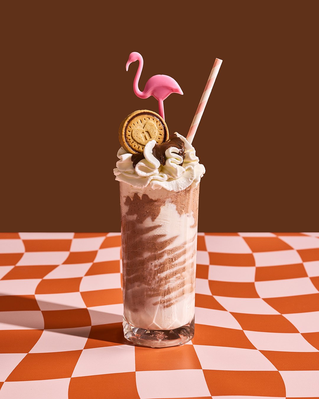 A milkshake in a tall glass with whipped cream, chocolate syrup, a cookie, a pink flamingo straw, and a clock-shaped decoration, all on an orange and white checkered surface with a plain brown background.