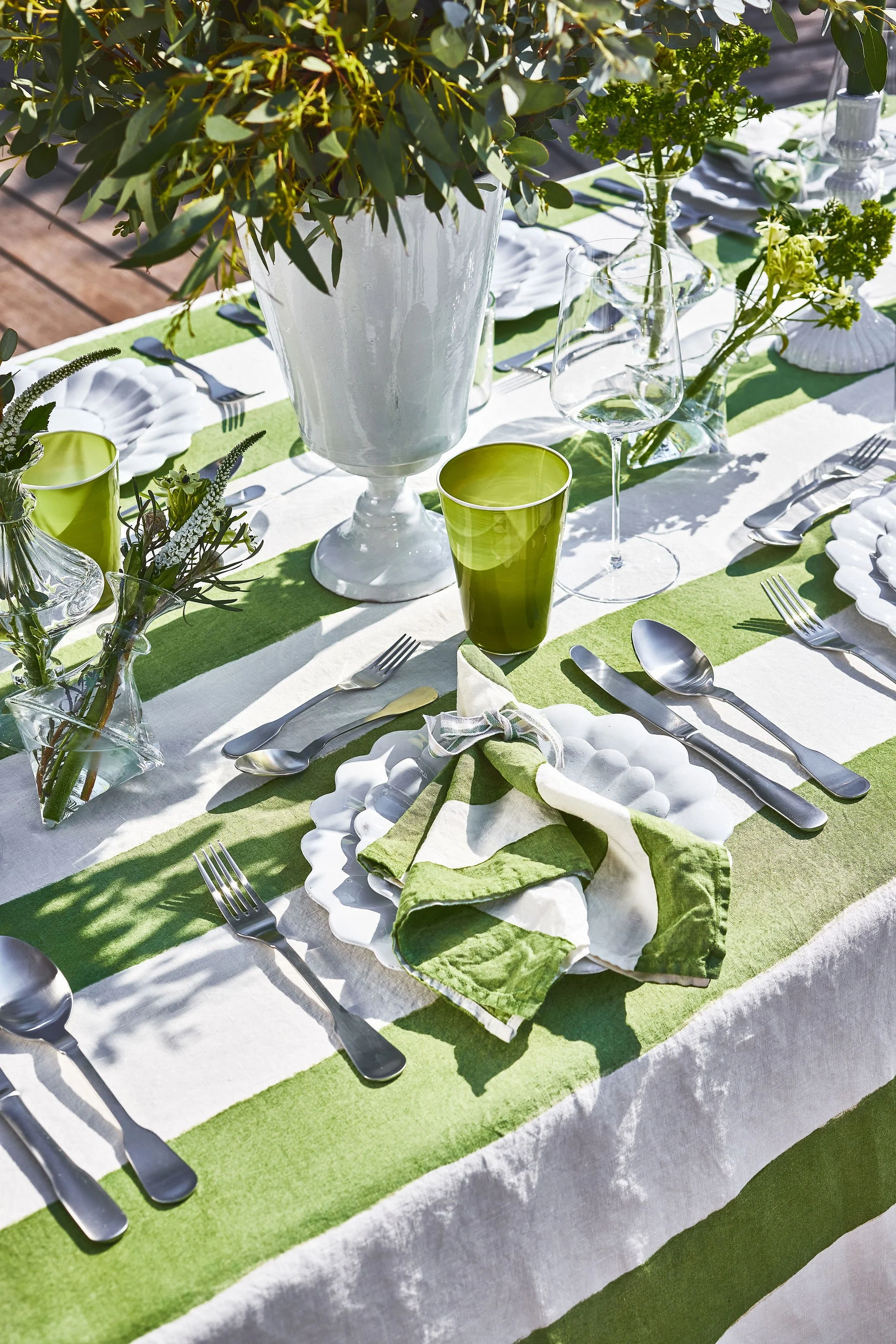 A table set outdoors with a white and green tablecloth, white plates, and silverware. There are green and clear glasses, and small vases with greenery. A large white vase with greenery is at the center.