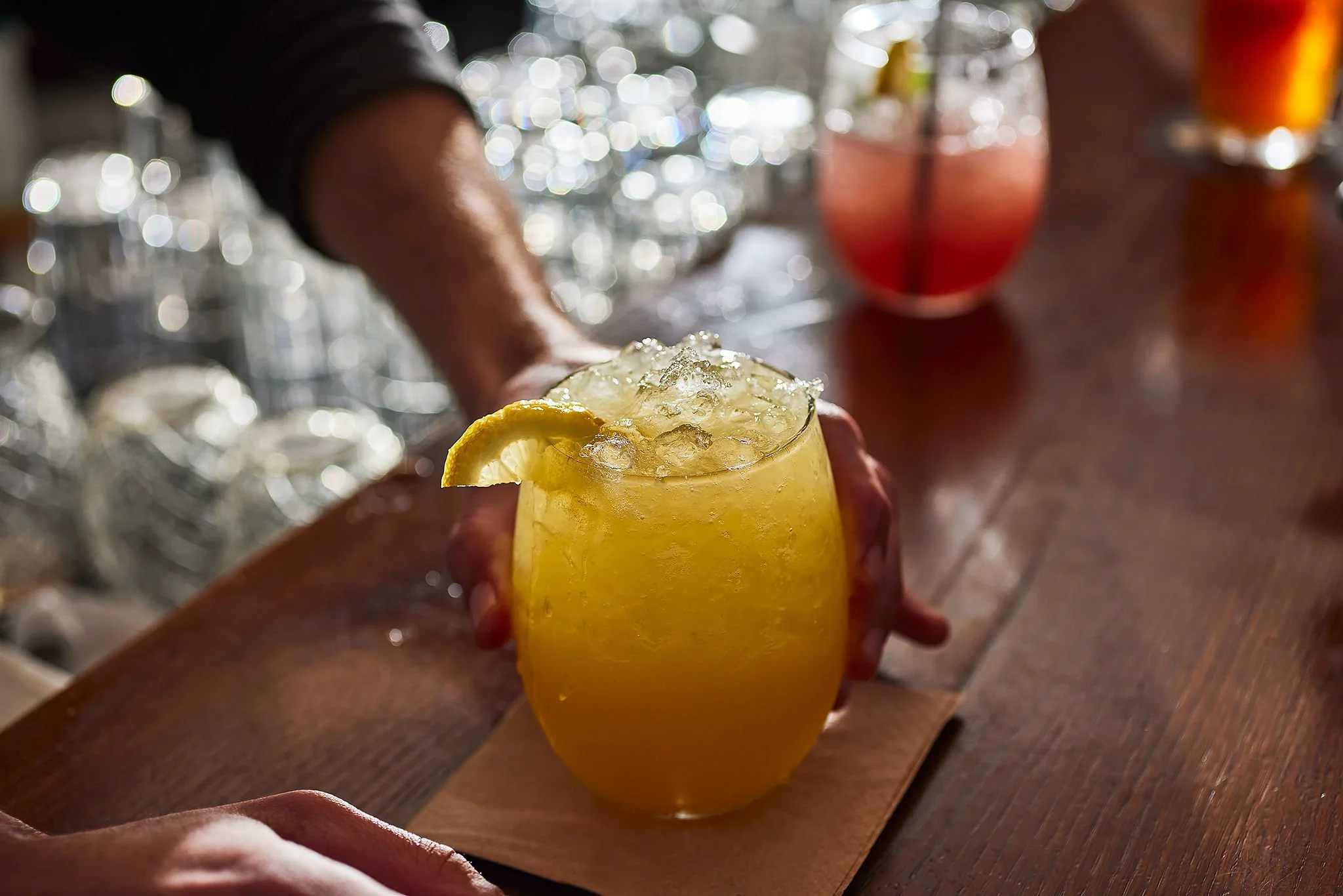 A person holding a yellow cocktail with ice and a lemon wedge garnish at a bar counter.