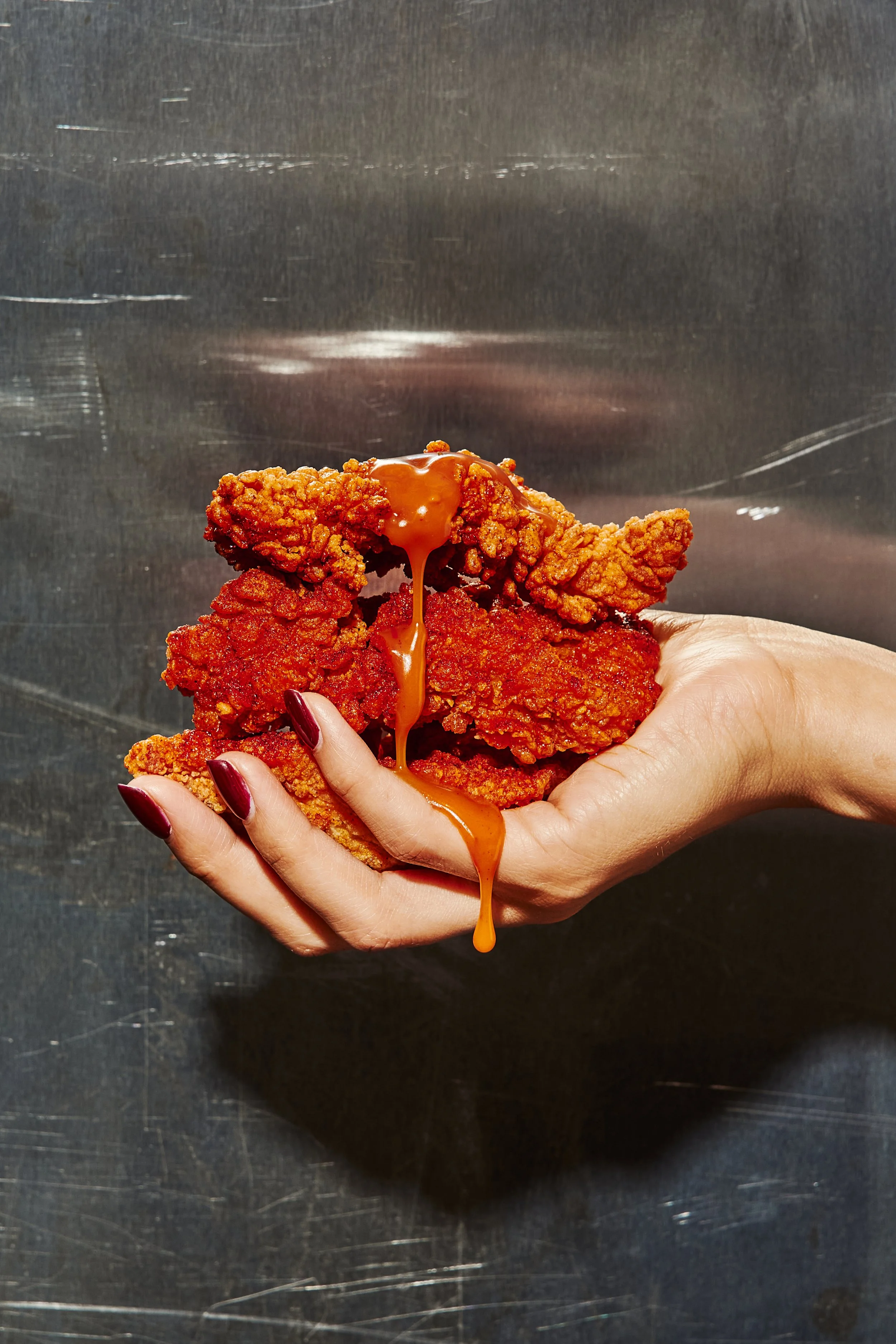 A person holding a pile of crispy fried chicken tenders with sauce dripping from them, against a metallic background. Tends kosher food in miami
