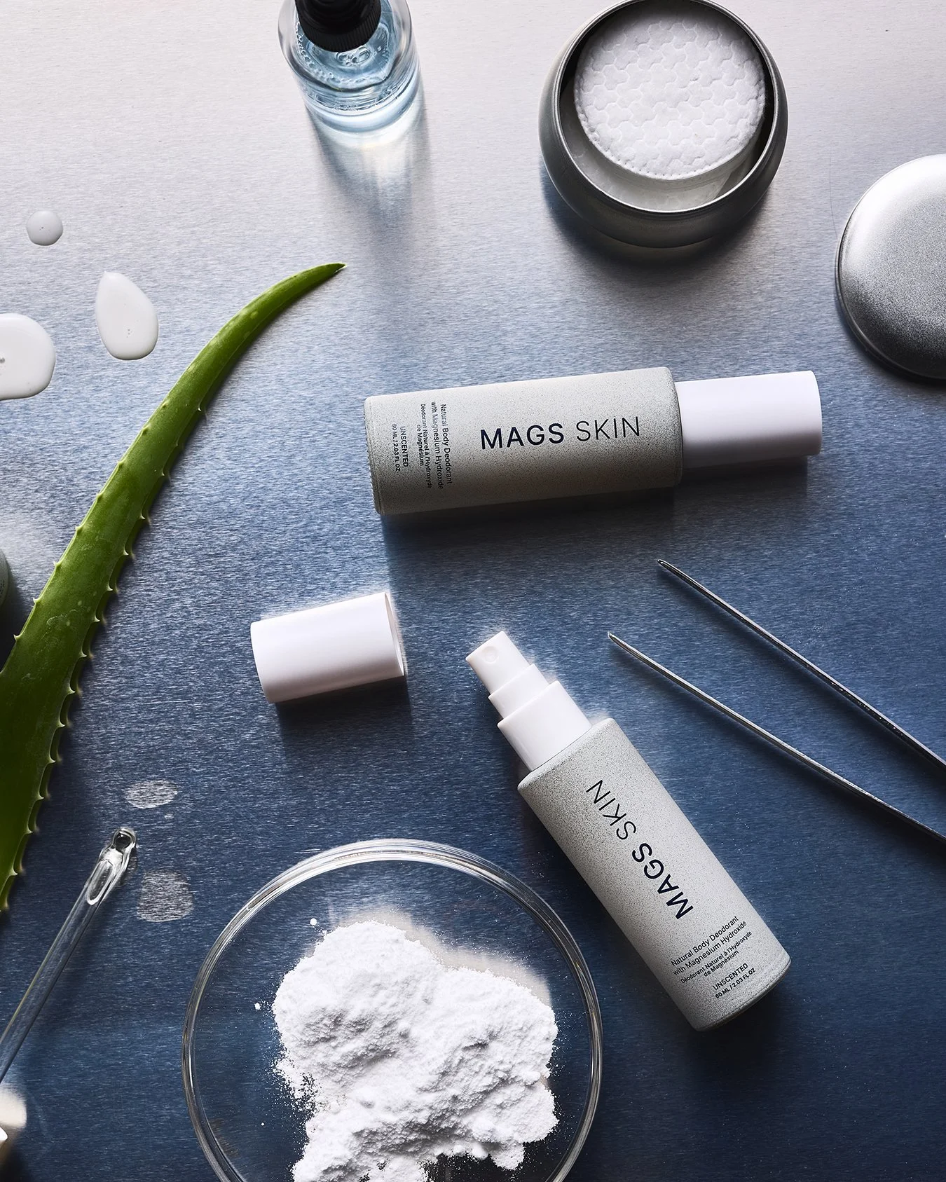 Skincare products on a blue surface, including MAGS SKIN moisturizer, a white spray bottle, a jar of powder, and a small glass bowl of white powder, along with aloe vera leaves, tweezers, a glass dropper, and a water bottle.