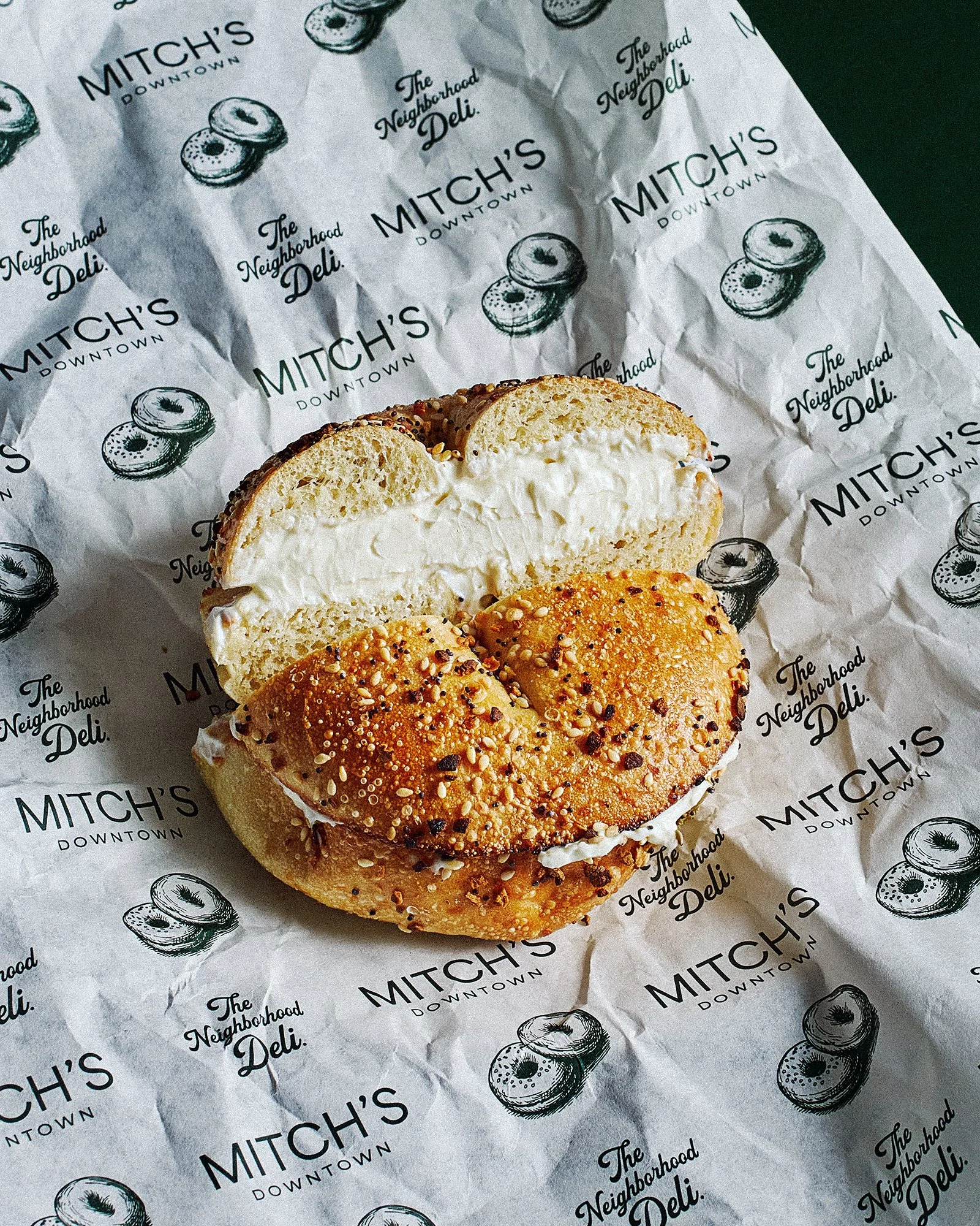 Bagel with cream cheese on crumpled paper with 'Mitch's Downtown' and 'The Neighborhood Deli' logos.