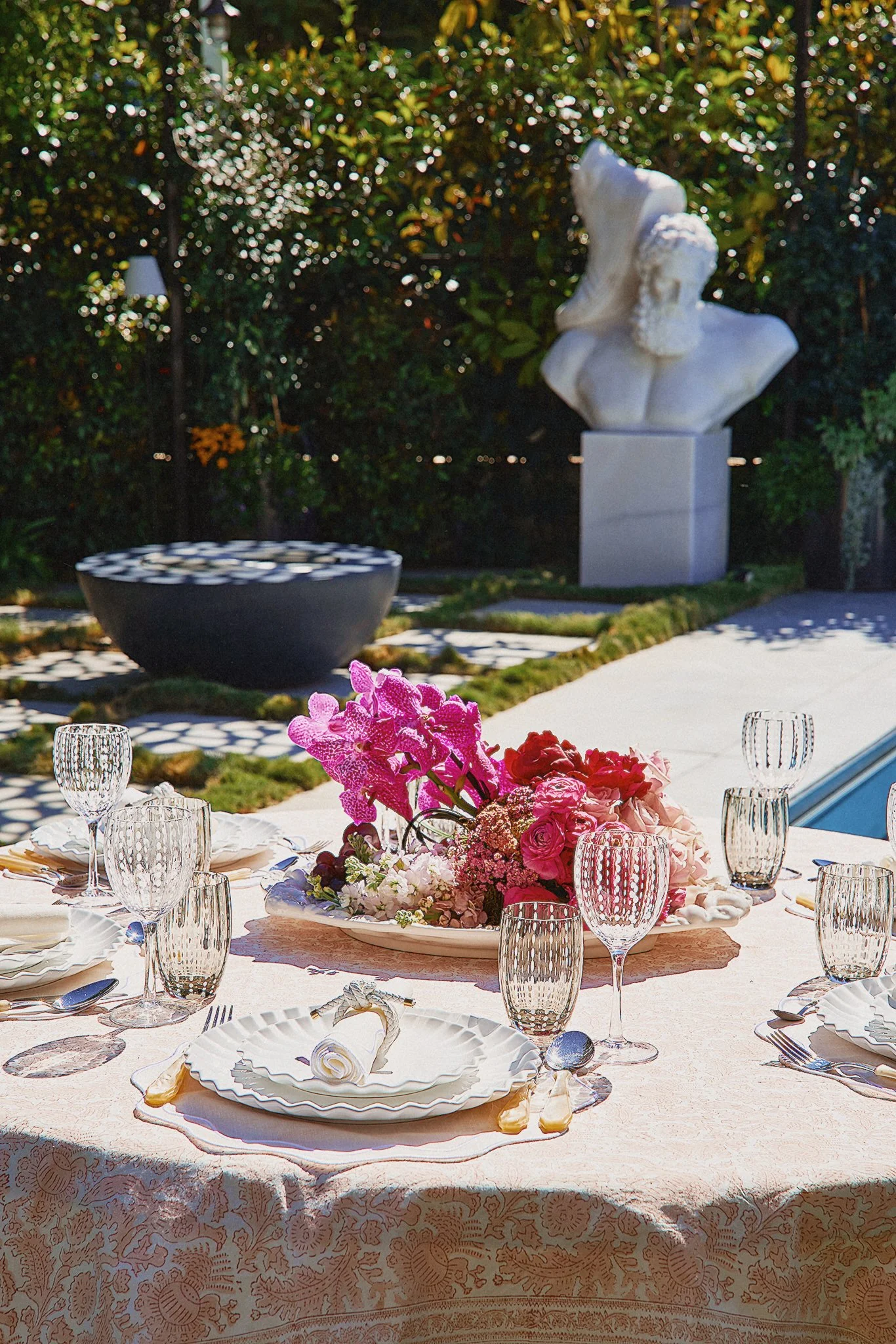 An outdoor dining table set with plates, glasses, and a floral centerpiece, with a garden and a white sculpture in the background.