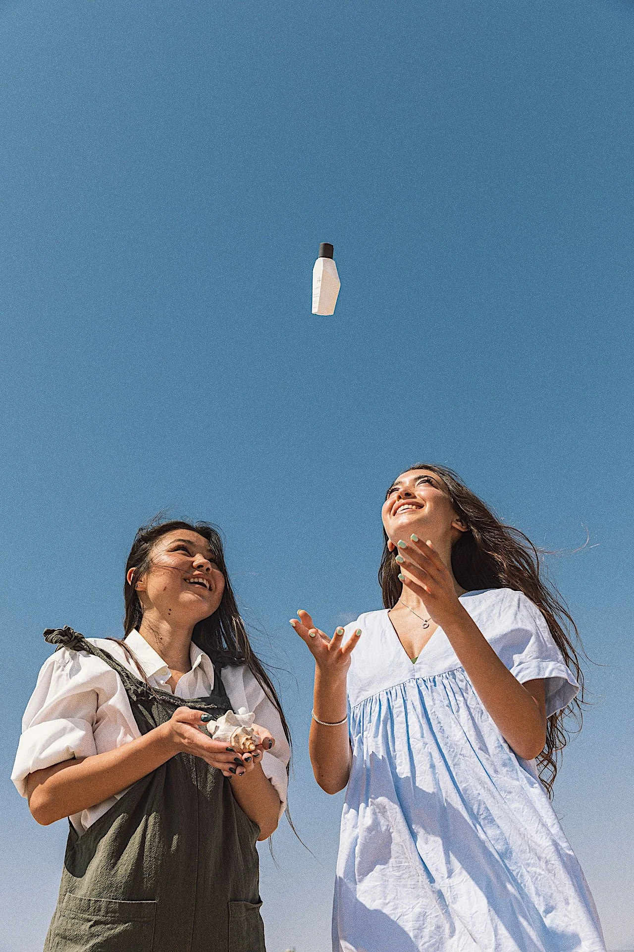 Two young women standing outdoors, looking up and smiling as a bottle is thrown into the air against a clear blue sky.