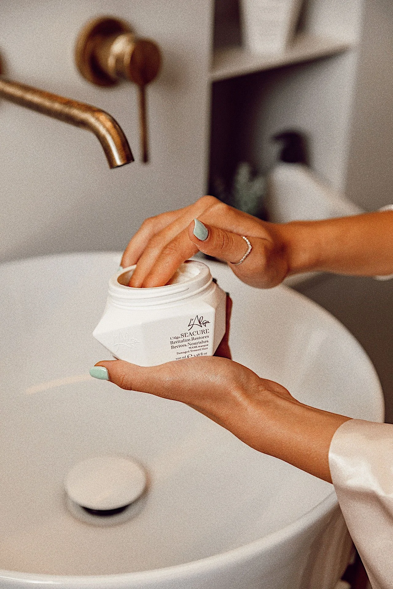 Person placing their finger into an open jar of skincare cream in a bathroom sink area.