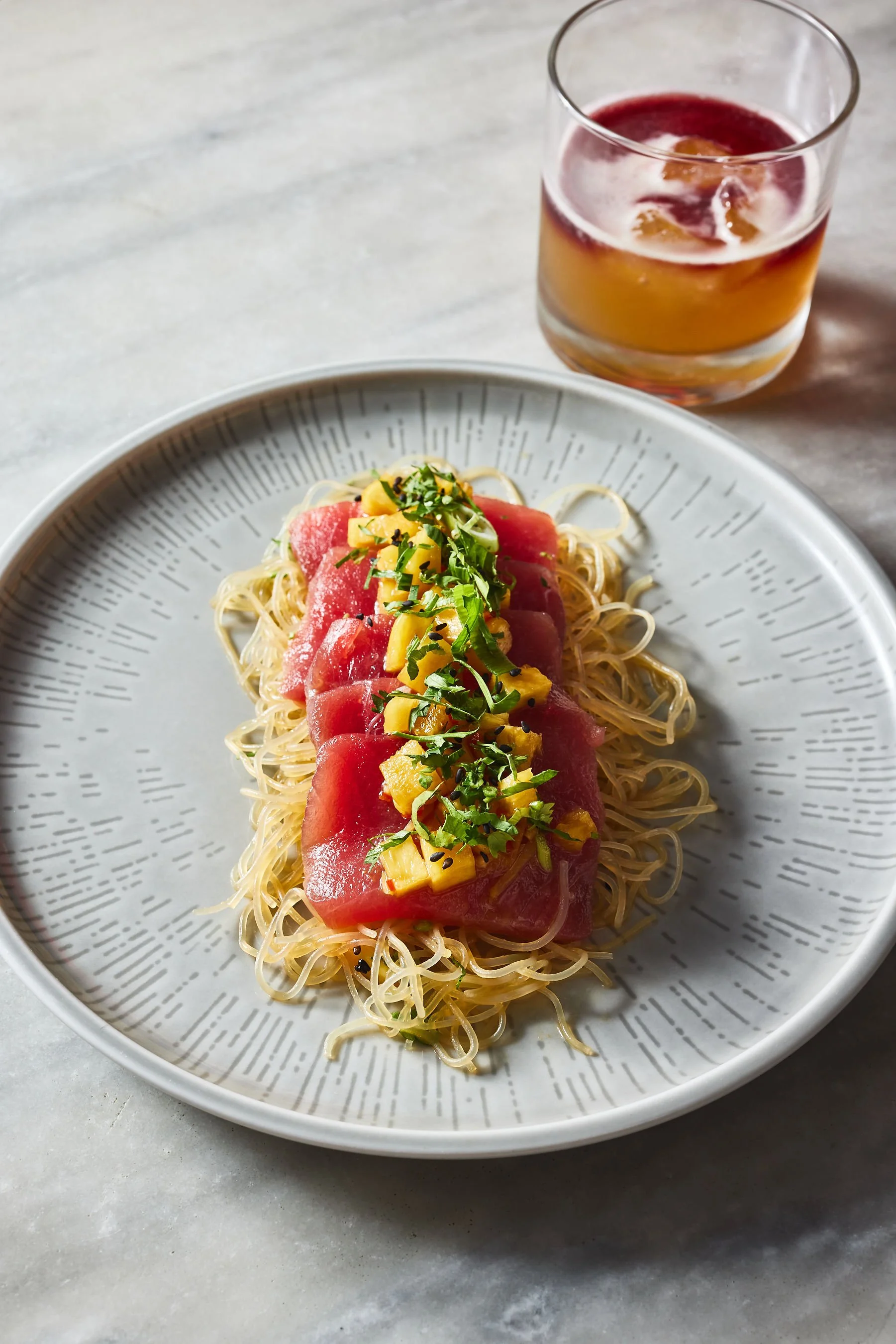 A plate of thin rice noodles topped with slices of watermelon, mango, and chopped herbs, served on a decorative gray plate, with a glass of red and yellow fruit juice on a light-colored surface.