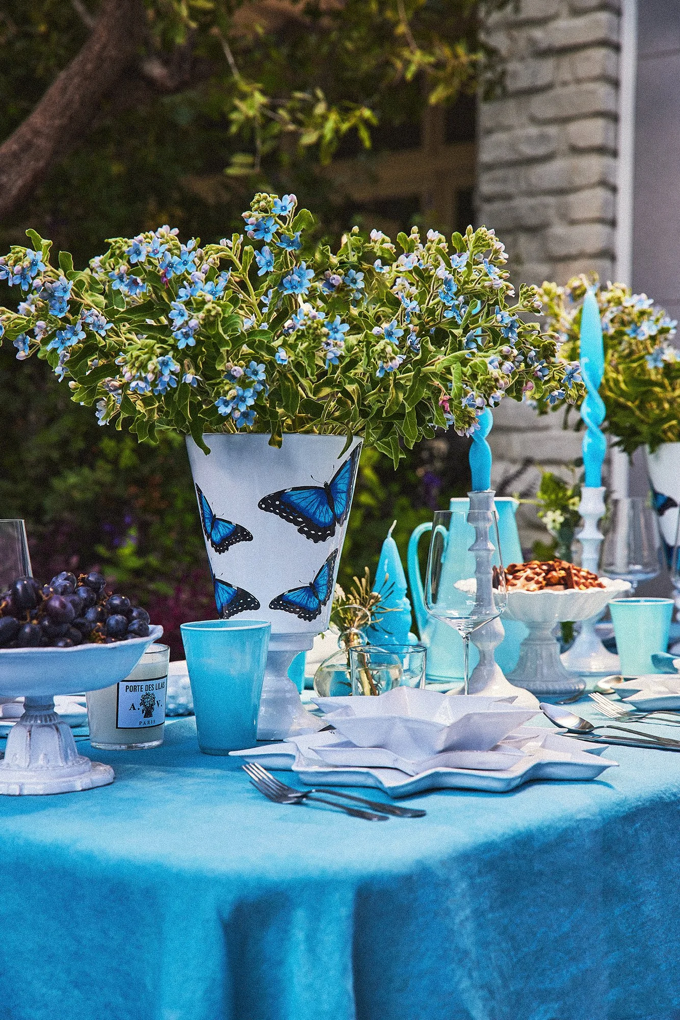 A decorated outdoor table with a blue tablecloth, featuring a large vase with blue butterfly designs filled with green and white flowering plants, surrounded by blue candles, bowls of grapes, dessert, glassware, and white napkins.