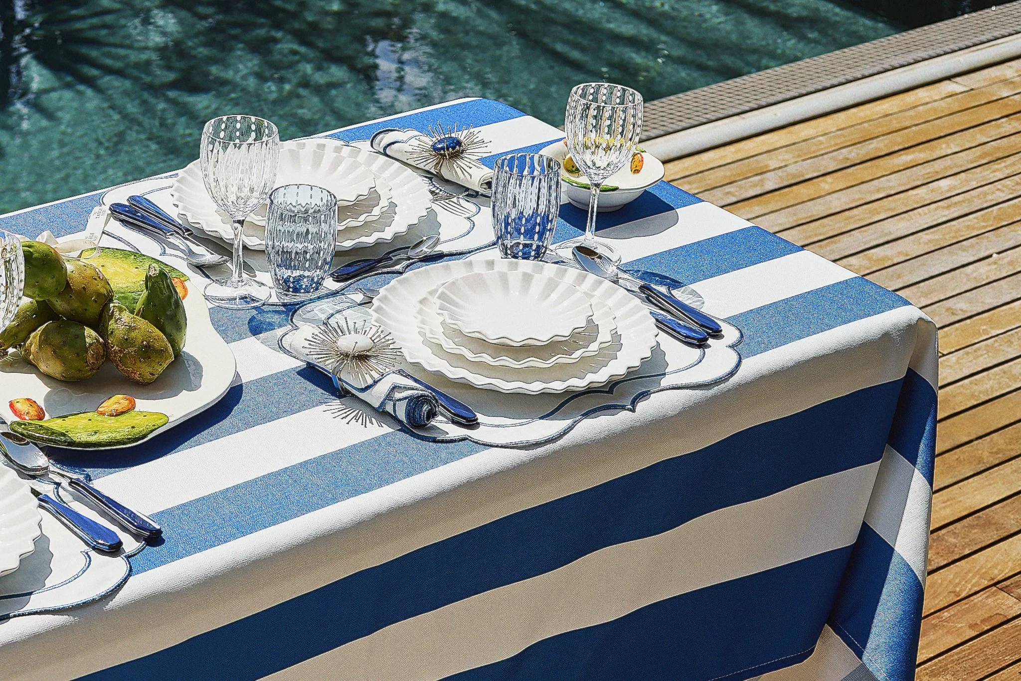 Table set for a meal with white, scalloped plates, water glasses, silverware, a salad with prickly pear cactus fruit, and blue and white striped tablecloth, near a pool with a wooden deck.