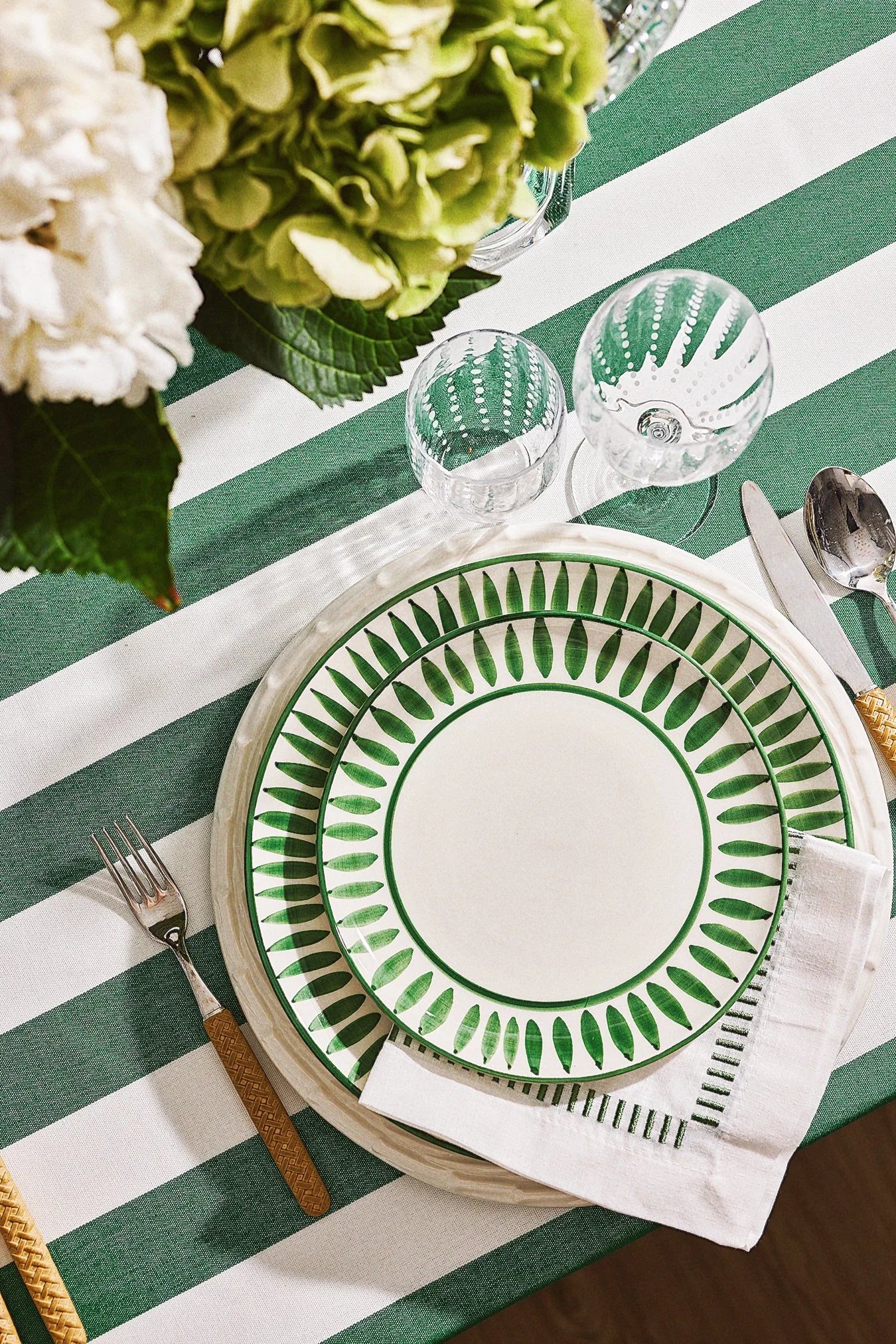 A table set for a meal with green and white striped tablecloth, green and white patterned plates, glassware, utensils, and a floral centerpiece with white and green flowers.
