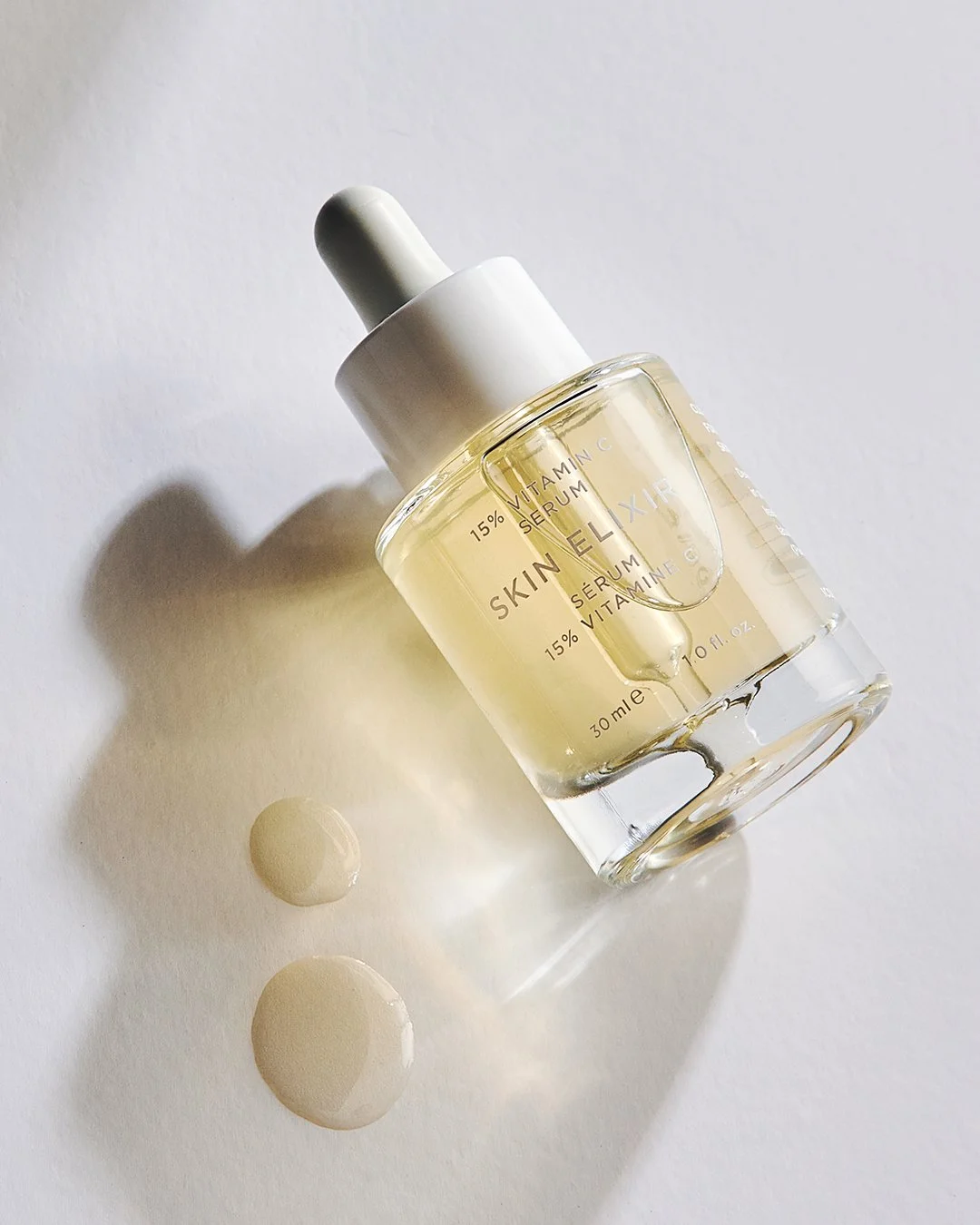 A clear glass bottle of Skin Elixir vitamin C serum with a white dropper cap, lying on a white surface with two drops of serum beside it.