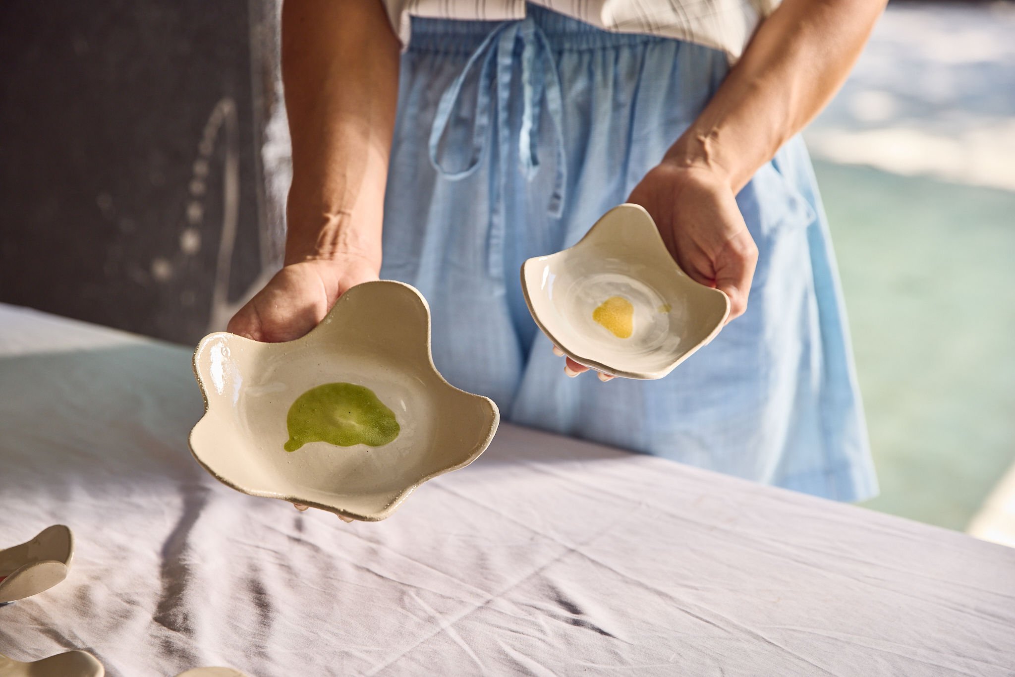 A person in light blue pajamas is holding two beige ceramic bowls with a scalloped edge, one with green sauce and the other with yellow sauce, over a white tablecloth with a body of water in the background.