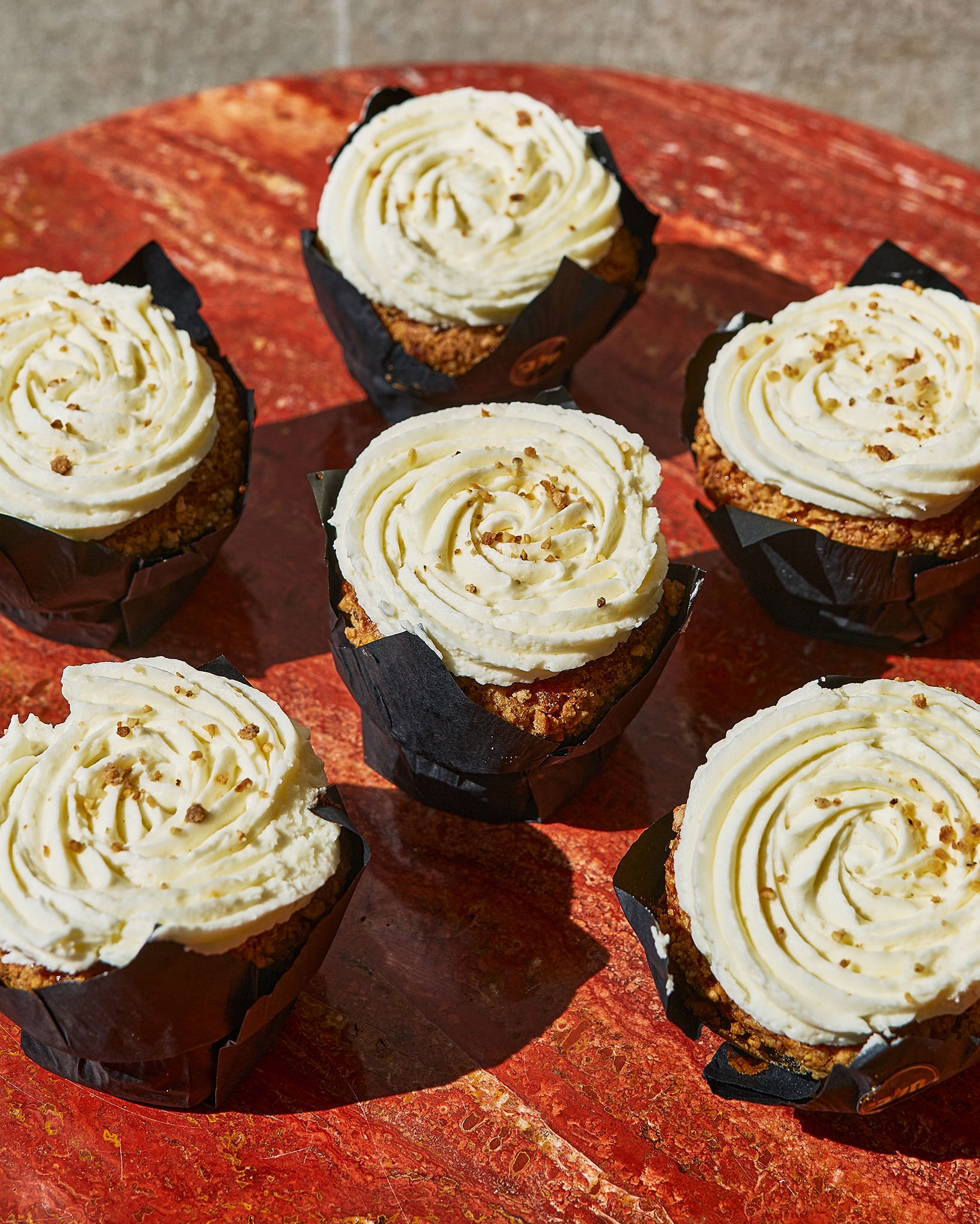 Six cupcakes with white frosting and sprinkles on a red surface. OREN MESHI