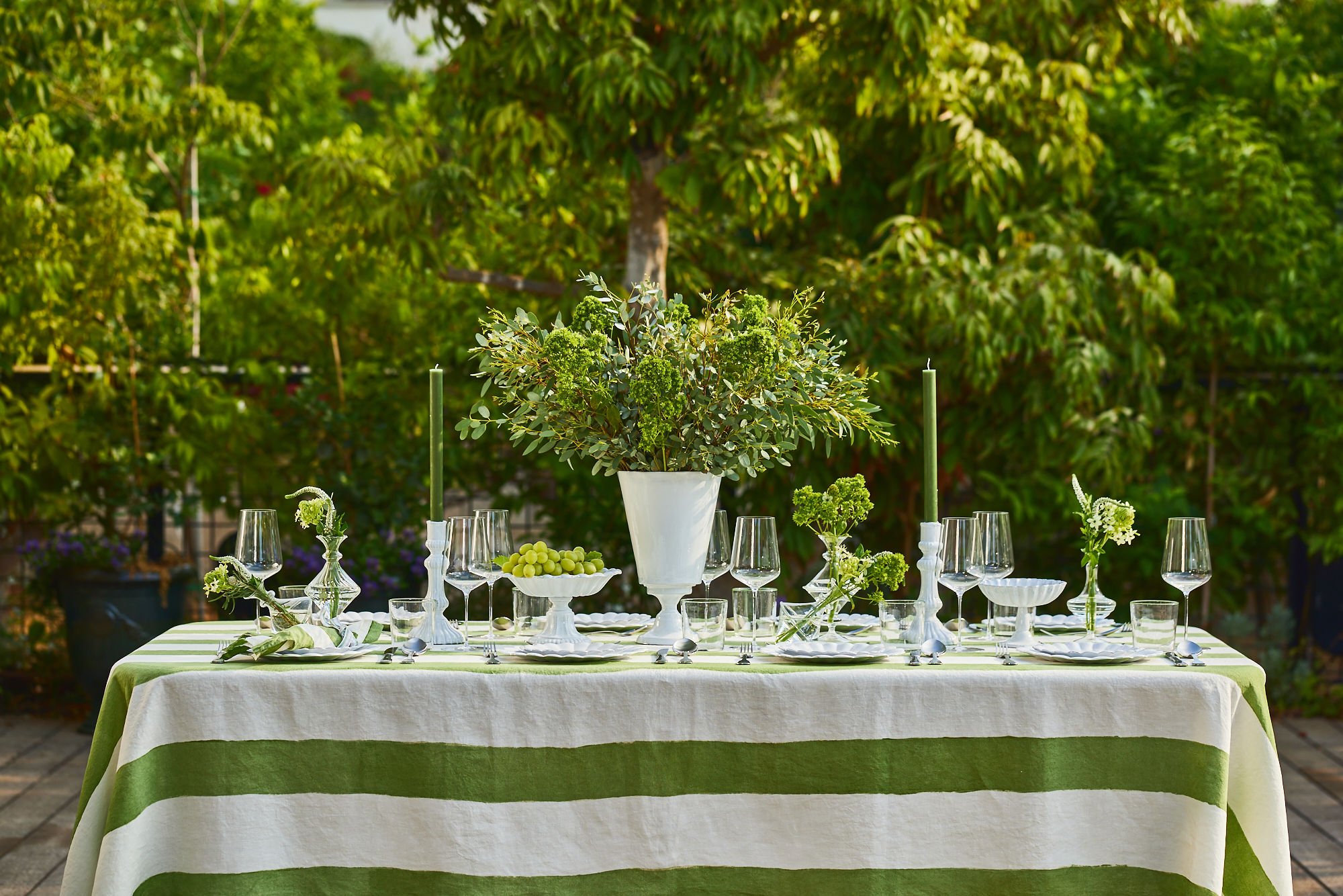 Outdoor dining table decorated with white and green striped tablecloth, glassware, white plates, green candles, and floral centerpieces, set in a lush garden setting.