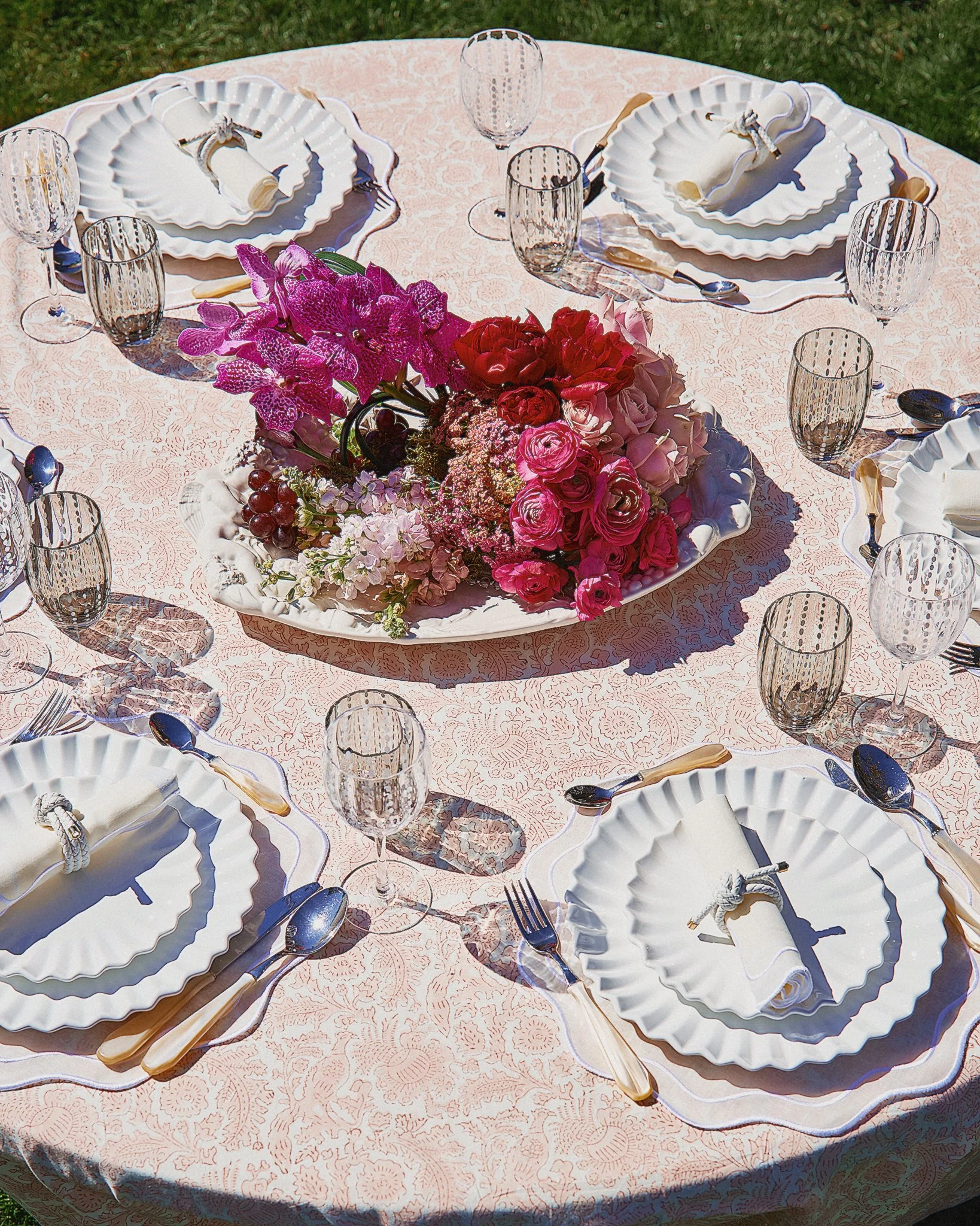 Round table set for a meal outdoors with a pink tablecloth, white plates, silverware, and empty glasses. A floral centerpiece with pink, red, purple, and white flowers is in the middle.