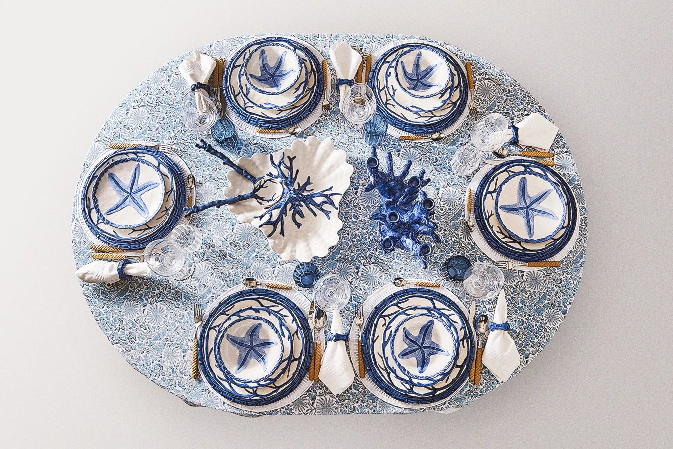 A nautical-themed table setting with plates, bowls, and glasses decorated with starfish and seaweed designs in blue and white, along with shell-shaped centerpieces and gold cutlery.
