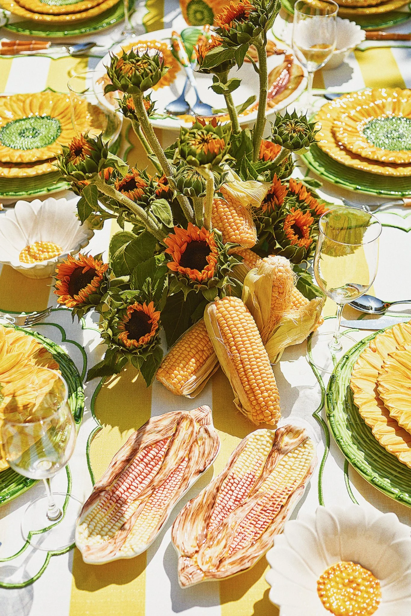Table centerpiece with sunflowers, corn on the cob, and autumn-themed decorations, surrounded by plates, glasses, and silverware for a meal.