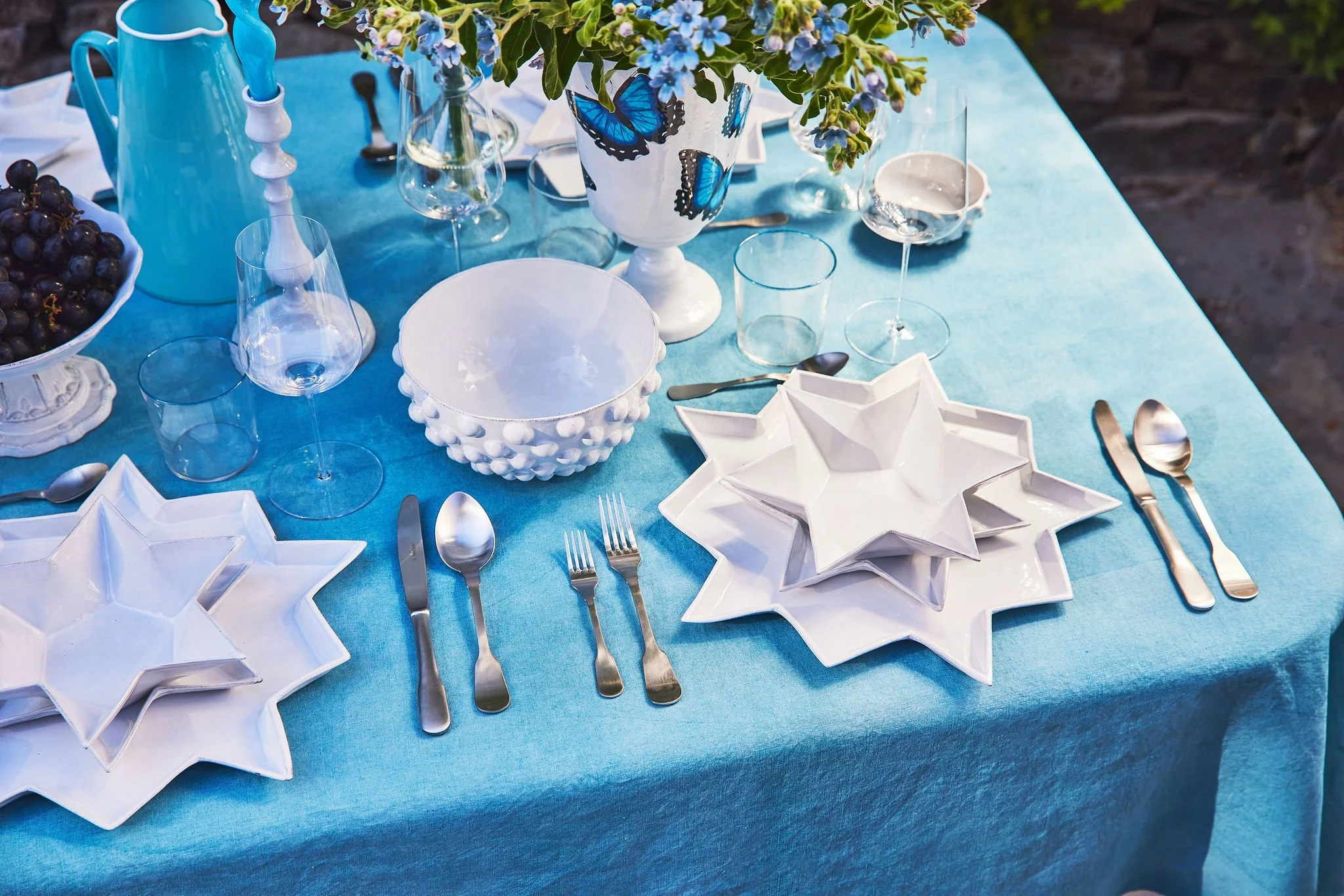 A beautifully set dining table with a bright blue tablecloth, white star-shaped dinnerware, various glassware, silver utensils, and a floral centerpiece with blue butterflies on a vase.