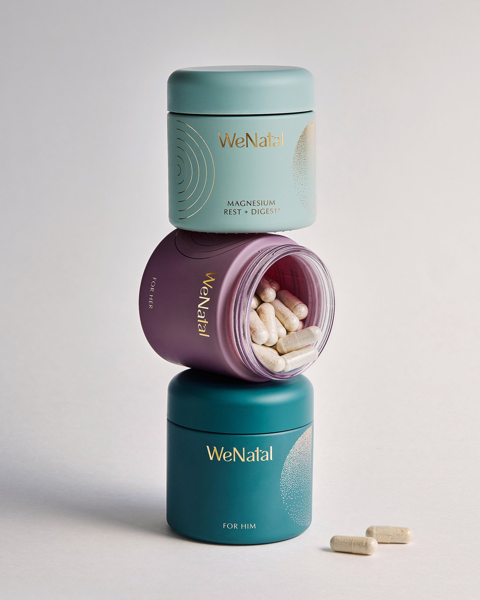 Stacked containers with medicine capsules, pink purple container open, pills spilling out, blue container on bottom labeled 'For Him', purple container in middle labeled 'For Her', top light blue container labeled 'Magnesium Rest + Digest'. WENATAL