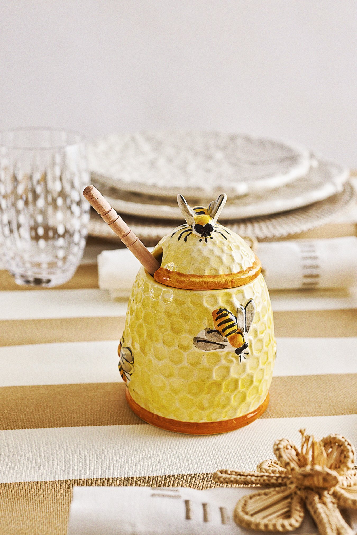 Decorative ceramic honey jar with bee illustrations and a bee figure on top, on a table with a striped tablecloth, surrounded by plates, glasses, and a starfish.
