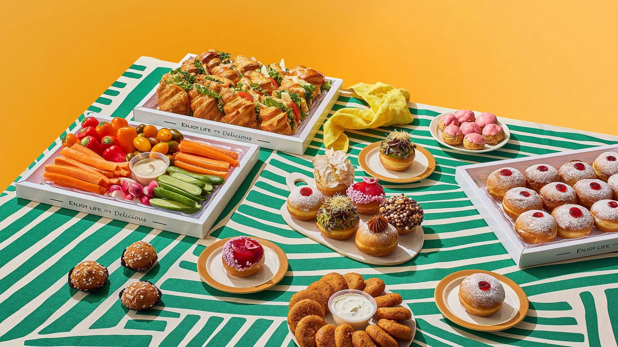 Fruit platter with cherry tomatoes, carrots, cucumbers, radishes, and dip; assorted sandwiches; cupcakes with pink and red frosting and toppings; assorted donuts with powdered sugar and cherry toppings; fried snacks with dipping sauce on a striped green and white tablecloth against a yellow background.