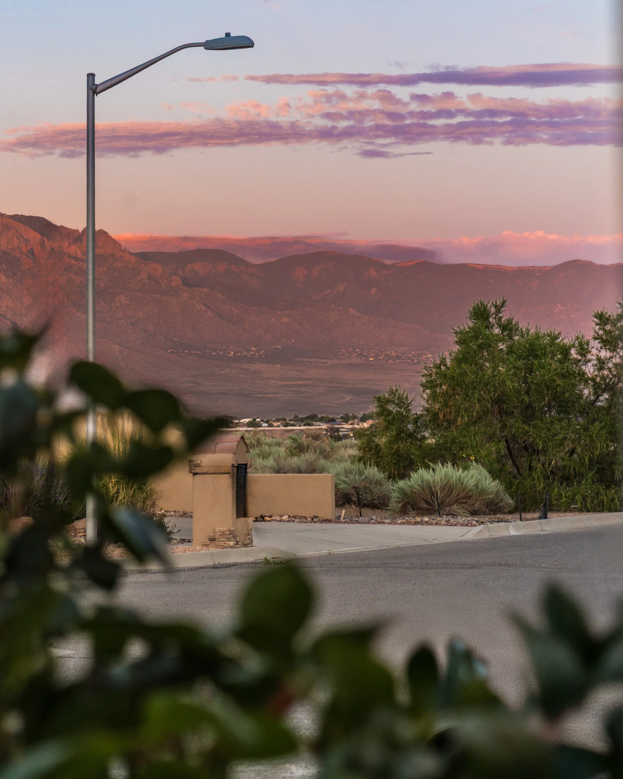 Street view with a street lamp, mountainous background, and a small building in the foreground, taken during sunset with purple and pink clouds in the sky.