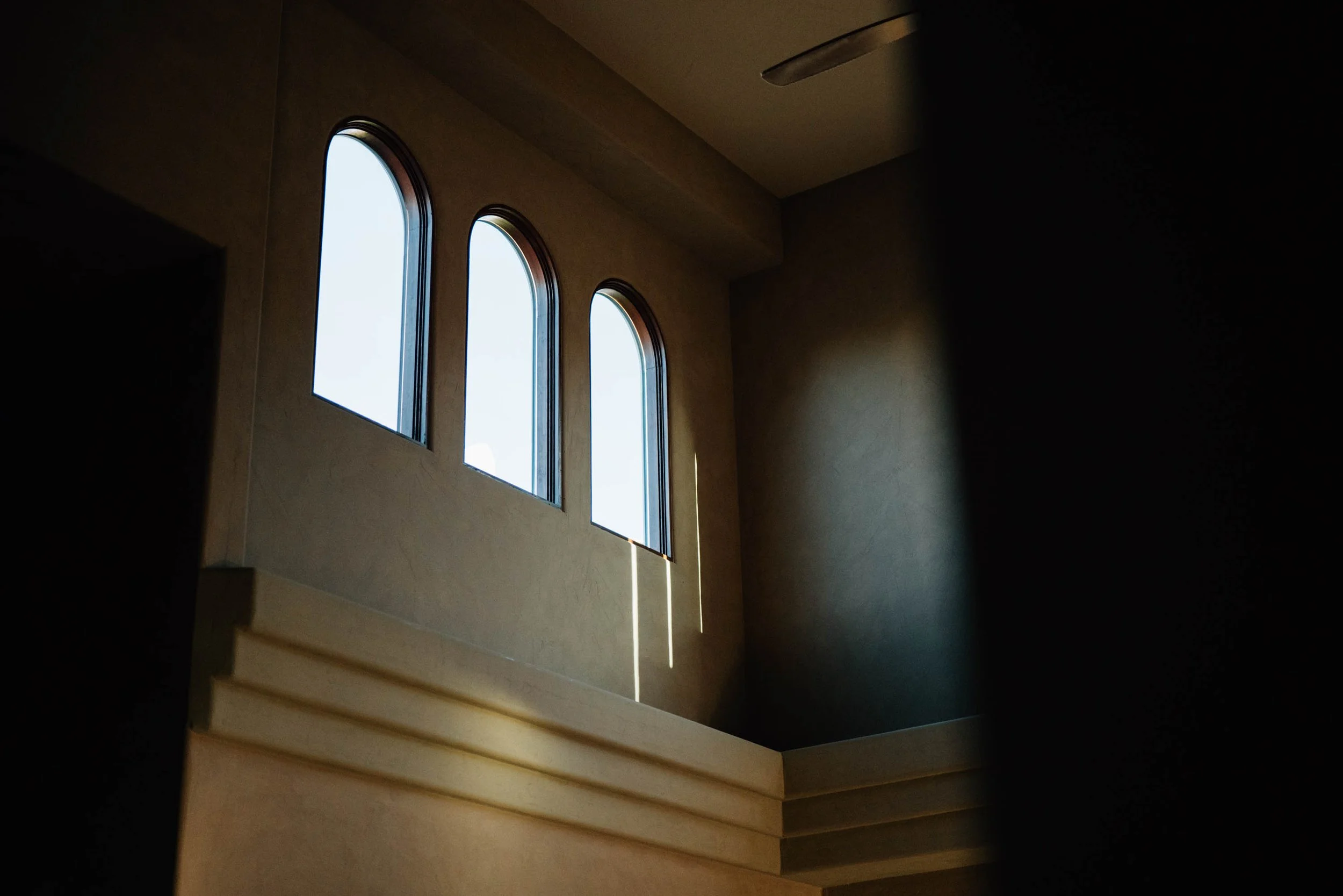 Interior view of a building with three tall, arched windows letting in sunlight, and wooden stairs on the right side.