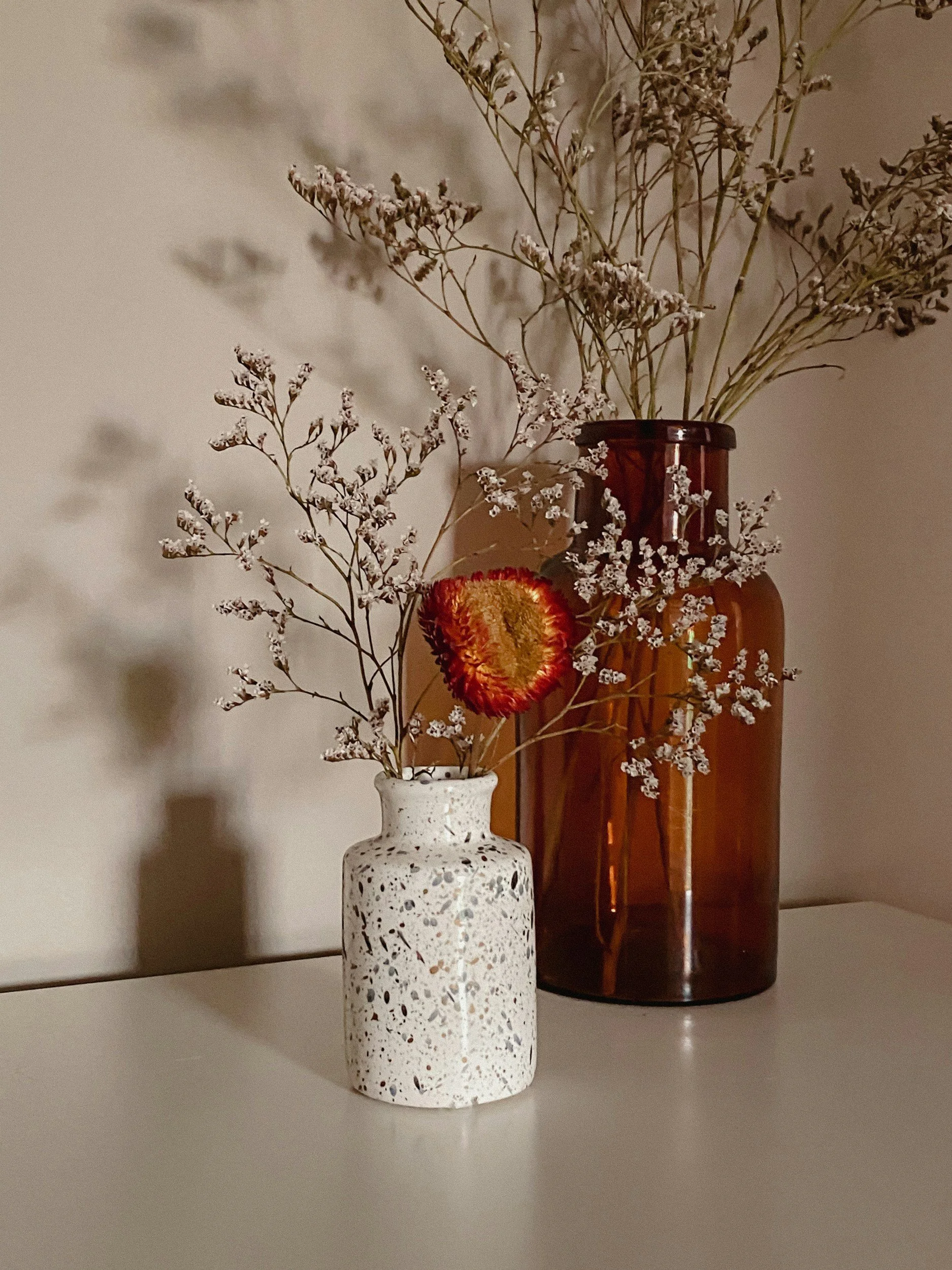 A still life of two vases with dried flowers placed on a white surface, with shadows cast on the wall behind.