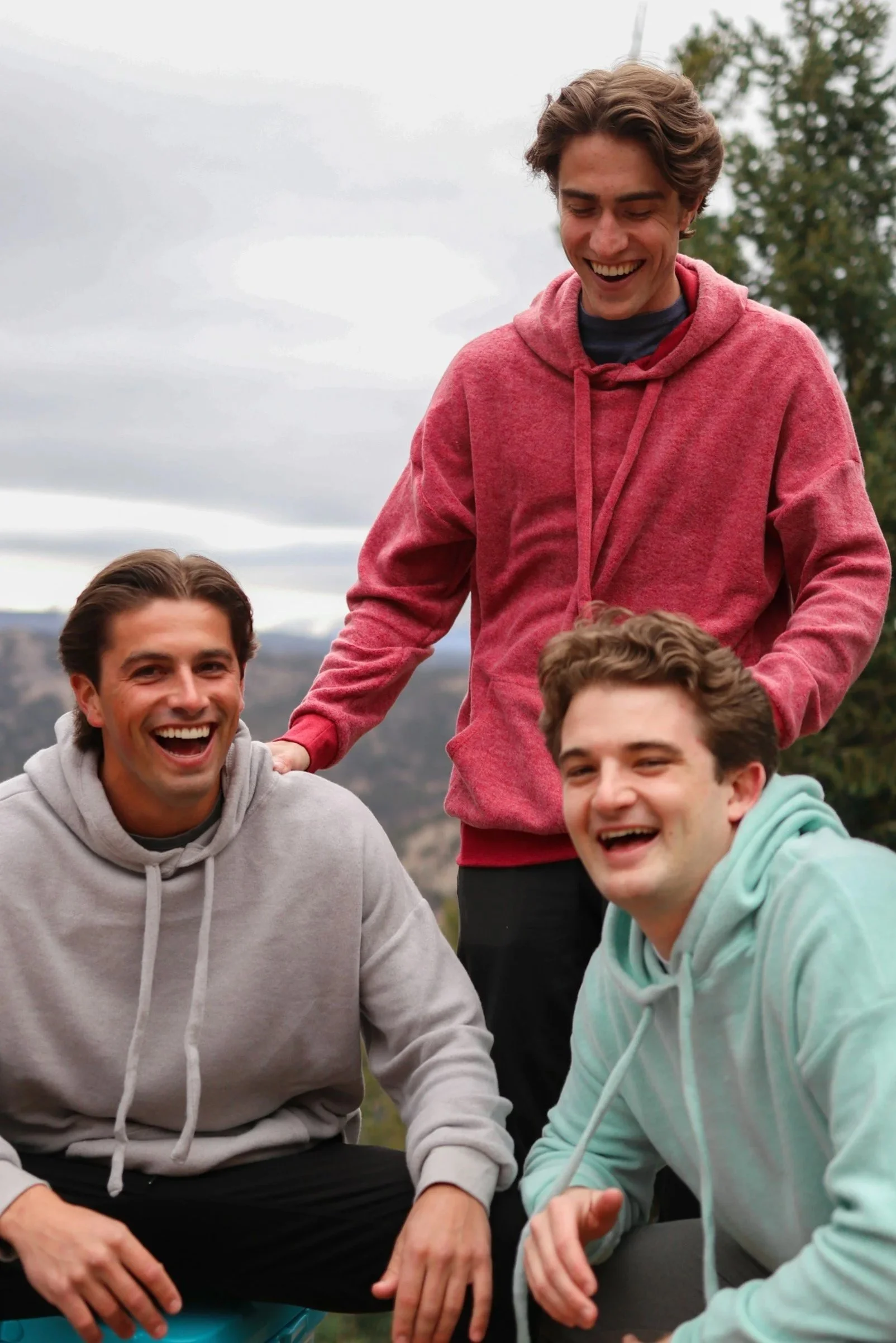 Four young men outdoors, smiling and laughing, dressed in casual hoodies, with a background of trees and cloudy sky.