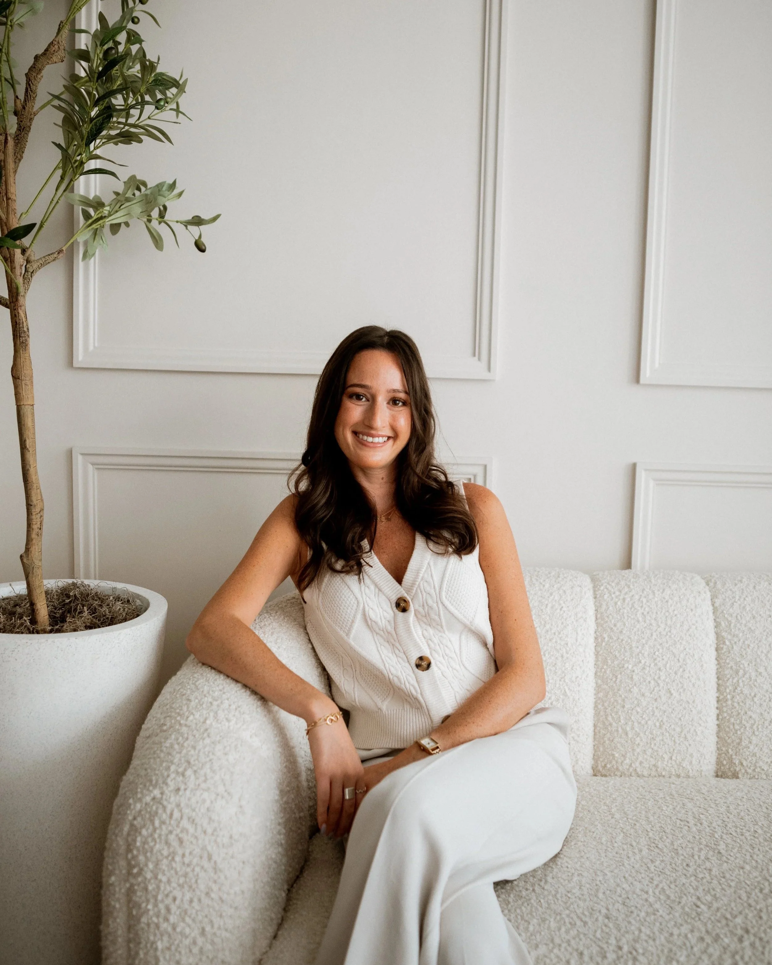A smiling woman with long brown hair sitting on a white textured sofa in a bright room with white walls and a large potted green plant.