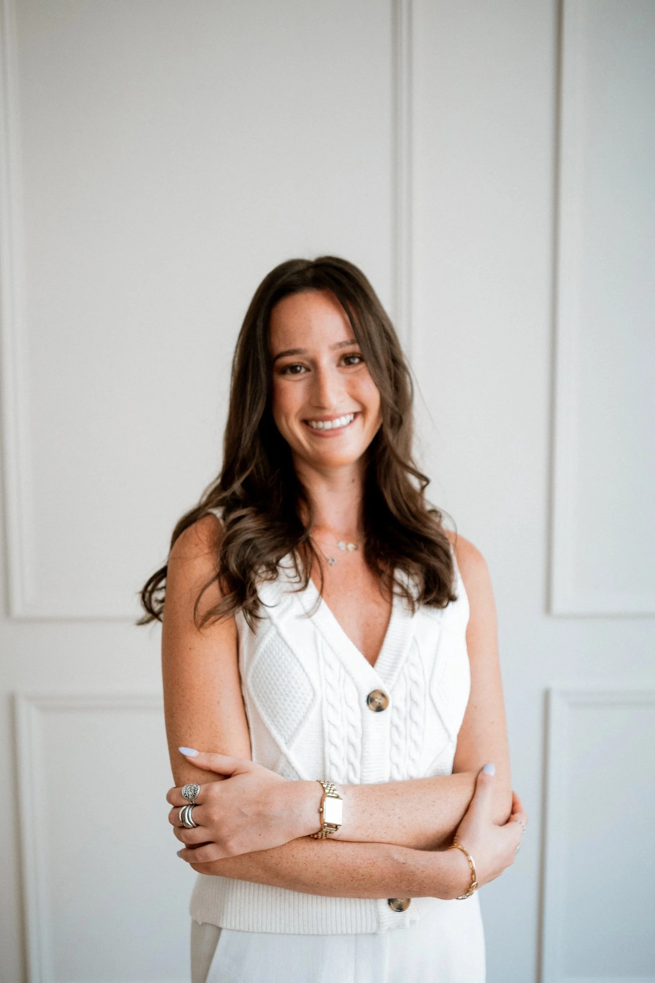 A woman with brown hair smiling and crossing her arms, wearing a white sleeveless knitted vest with buttons, jewelry including rings, a bracelet, and a watch, standing in front of white paneled walls.