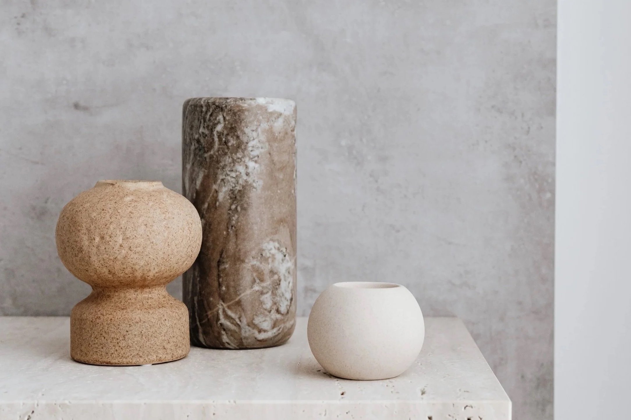Three minimalist vases with various textures and colors on a white surface against a gray wall.