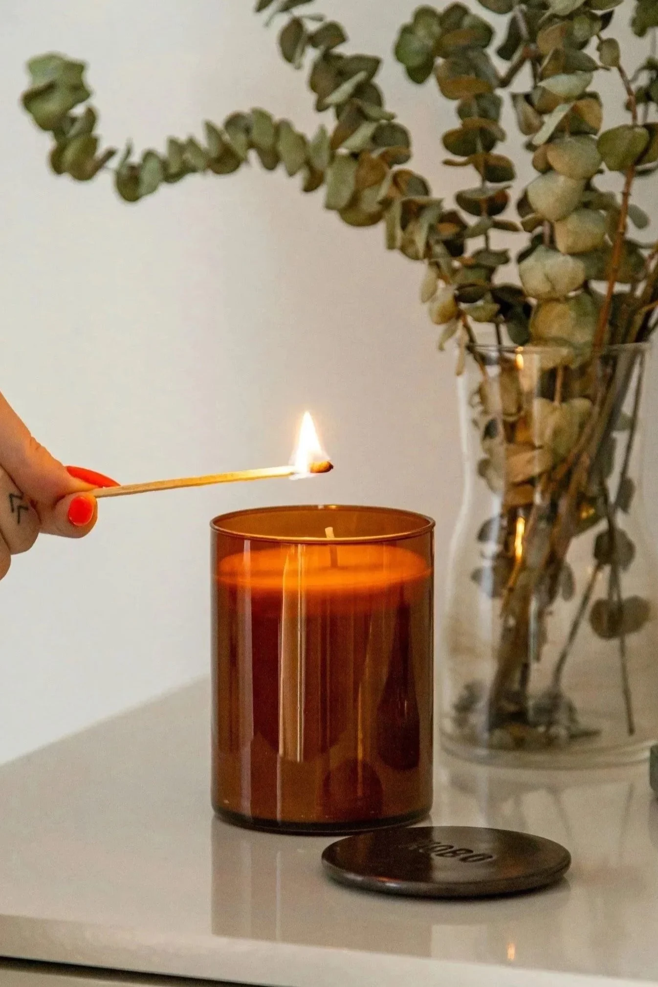 A person with painted nails holding a lit matchstick over an orange candle on a white surface, with a glass vase containing dried eucalyptus leaves in the background.
