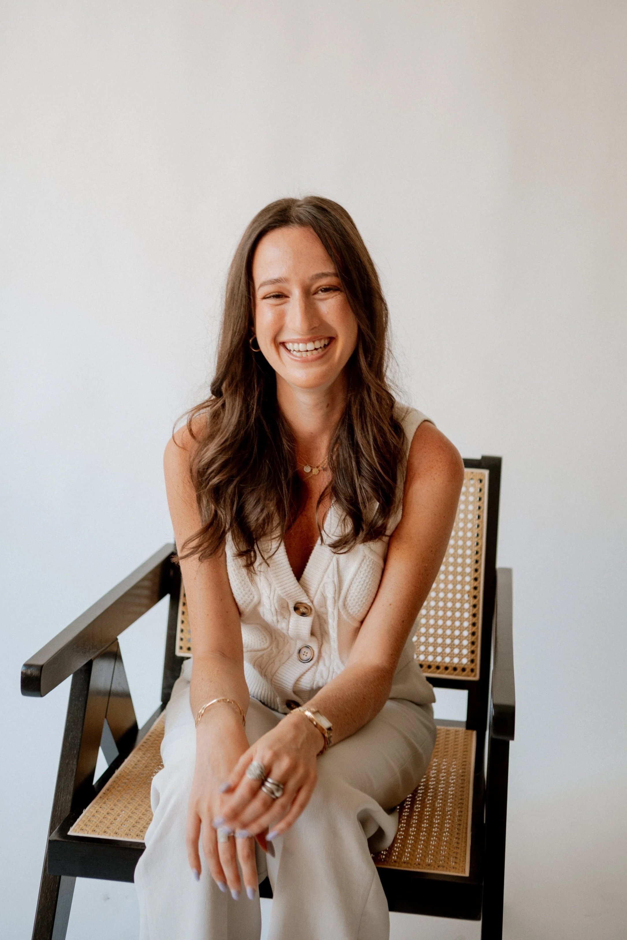 A woman with long, wavy brown hair smiling and seated on a black and tan cane-back chair against a plain white background. She is wearing a sleeveless cream-colored vest with buttons and several pieces of jewelry, including rings, bracelets, and a delicate necklace.
