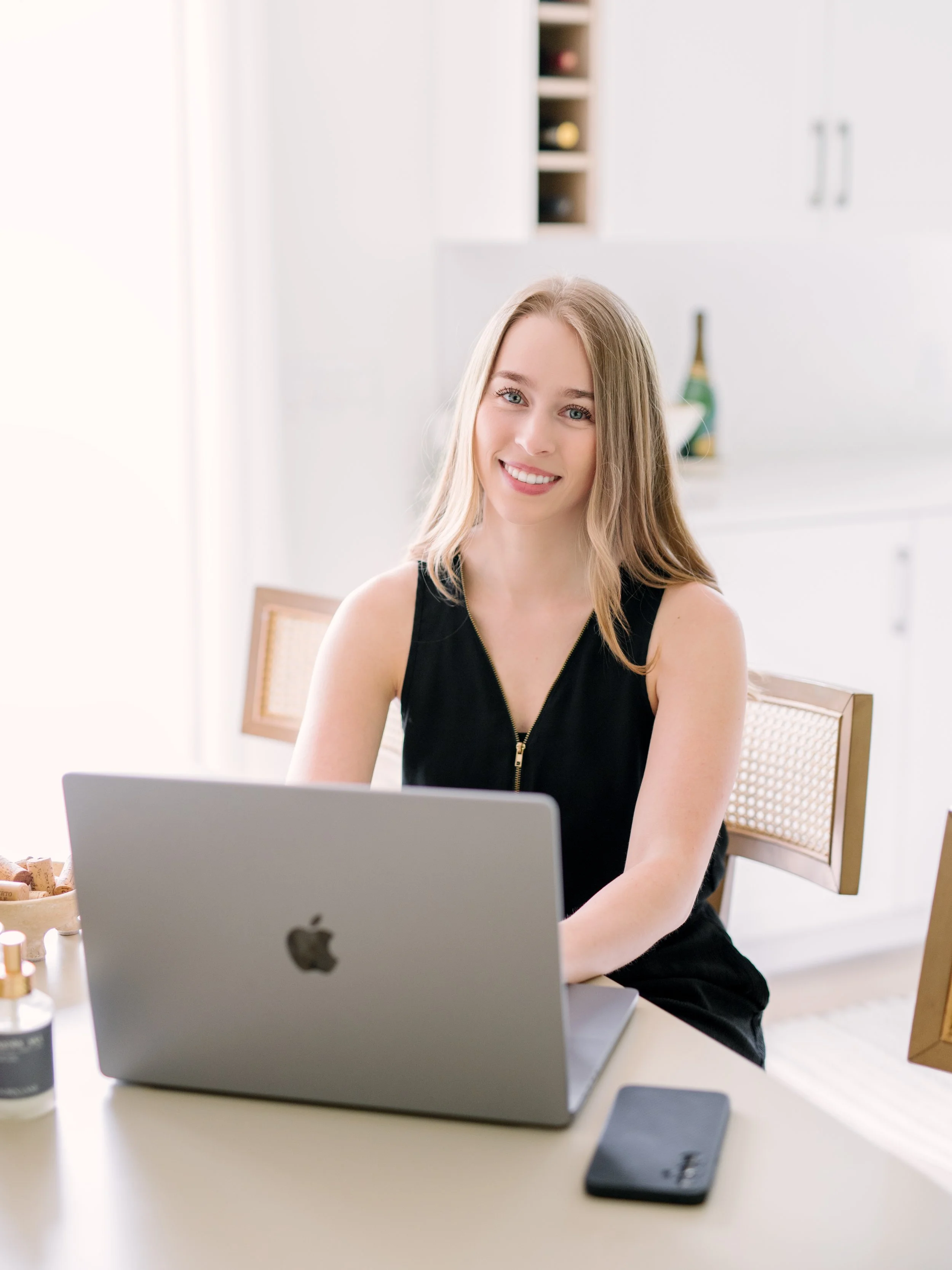 A young woman with blonde hair, wearing a black sleeveless top, sitting at a white table with a silver Apple MacBook in front of her, smiling at the camera in a bright, modern kitchen.