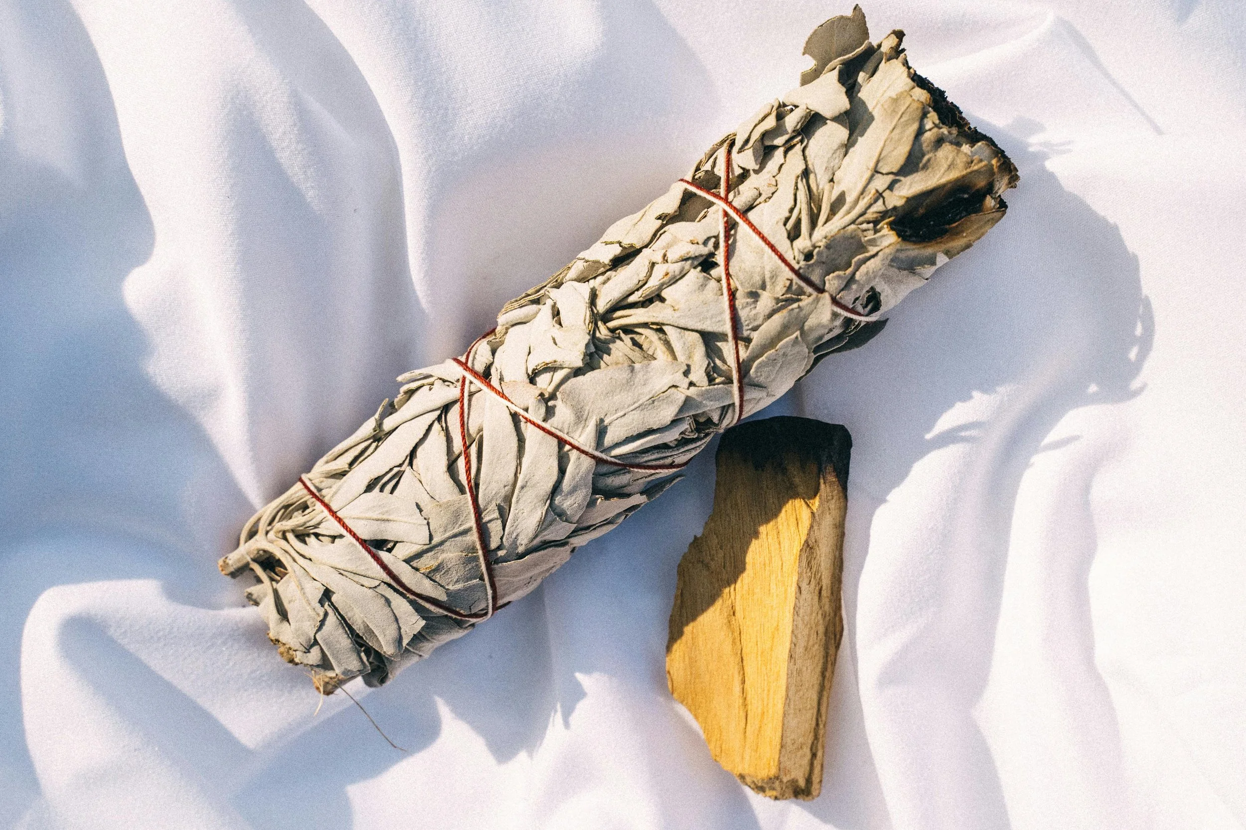 A bundle of white sage wrapped with red string and a piece of yellowish sage stick on white cloth.