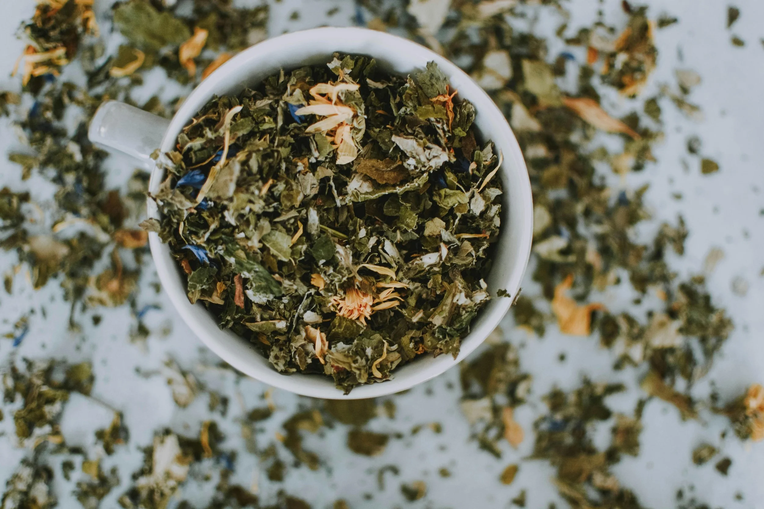 Top-down view of a white mug filled with loose herbal tea leaves and dried flowers, with scattered tea leaves and flowers on a light surface.