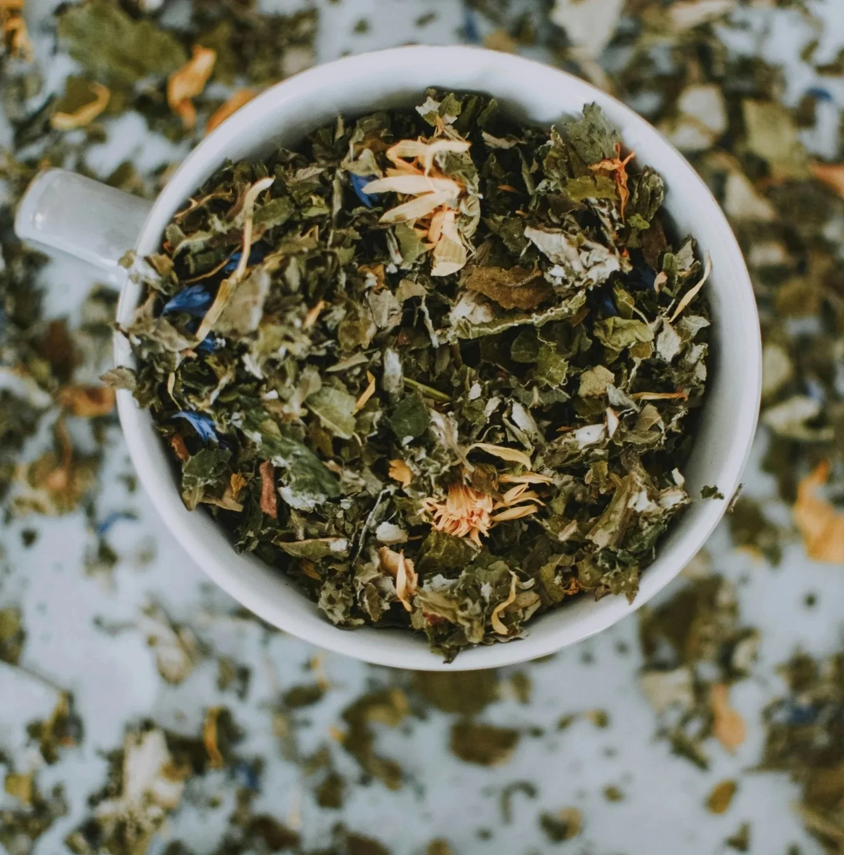 A white mug filled with dried herbal tea leaves and flower petals, with loose tea leaves scattered around on a surface.