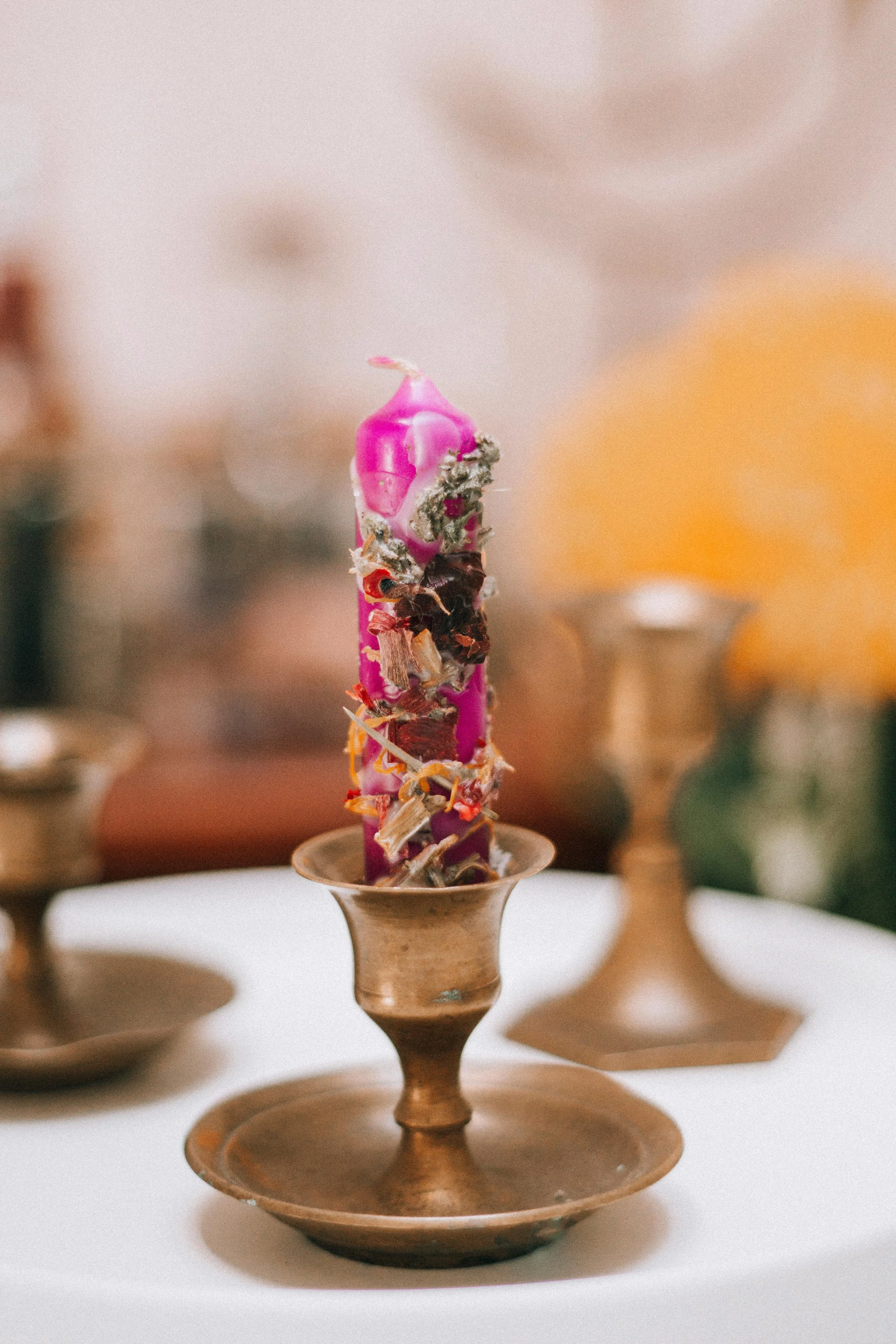 A purple birthday candle appears to be melted and surrounded by dried flower petals or potpourri on a brass candlestick holder, with a blurred background.