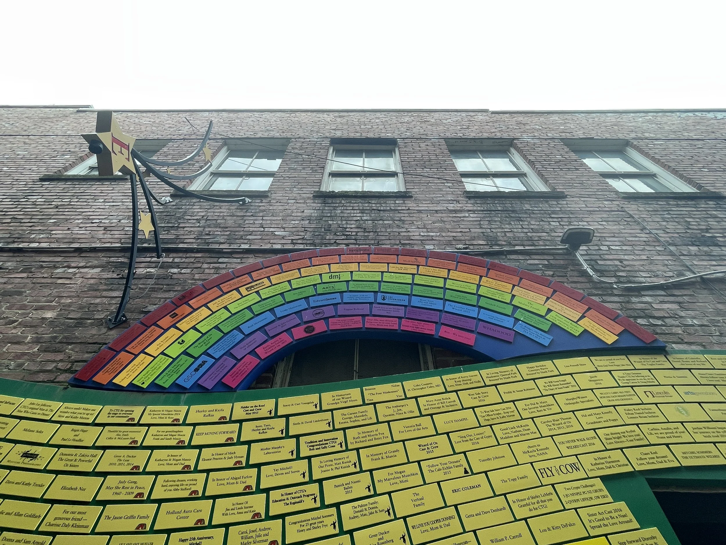 Colorful memorial wall with yellow, pink, purple, green, blue, and orange plaques mounted on a brick building exterior with four windows and a star-shaped decorative sign, between the stars and the yellow plaques is a rainbow.