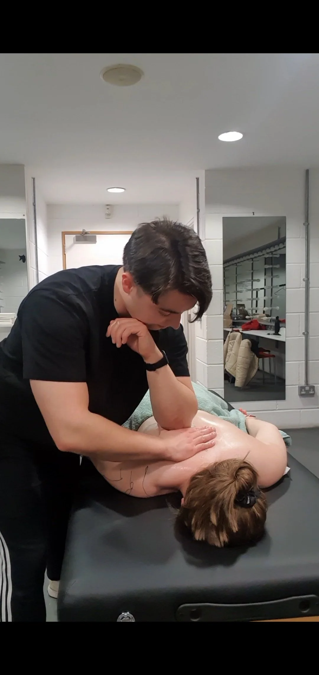 A person giving a massage to a woman lying face down on a massage table in a room with white walls and ceiling lights.