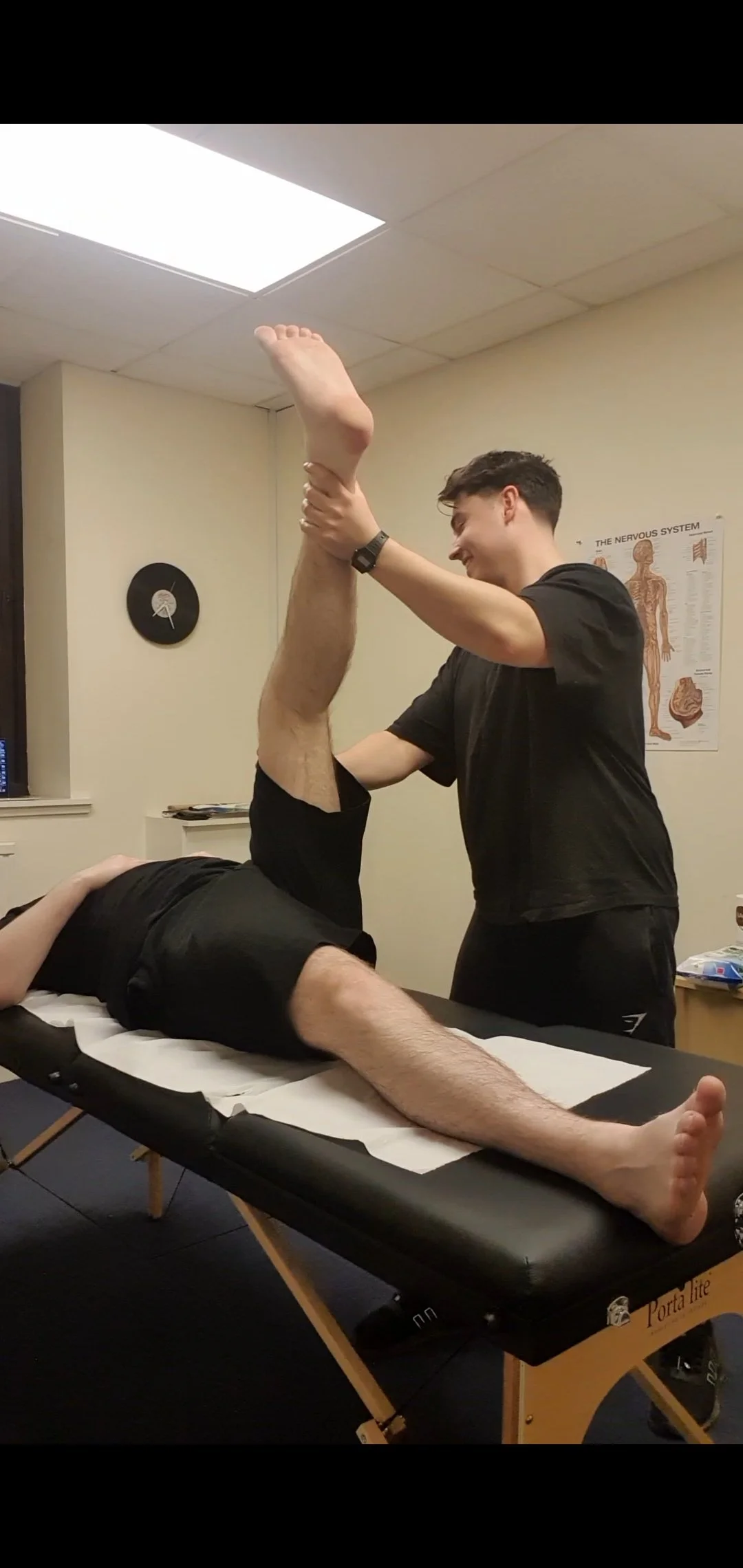 A physical therapist or chiropractor assisting a patient with leg stretches on a treatment table in a medical office.