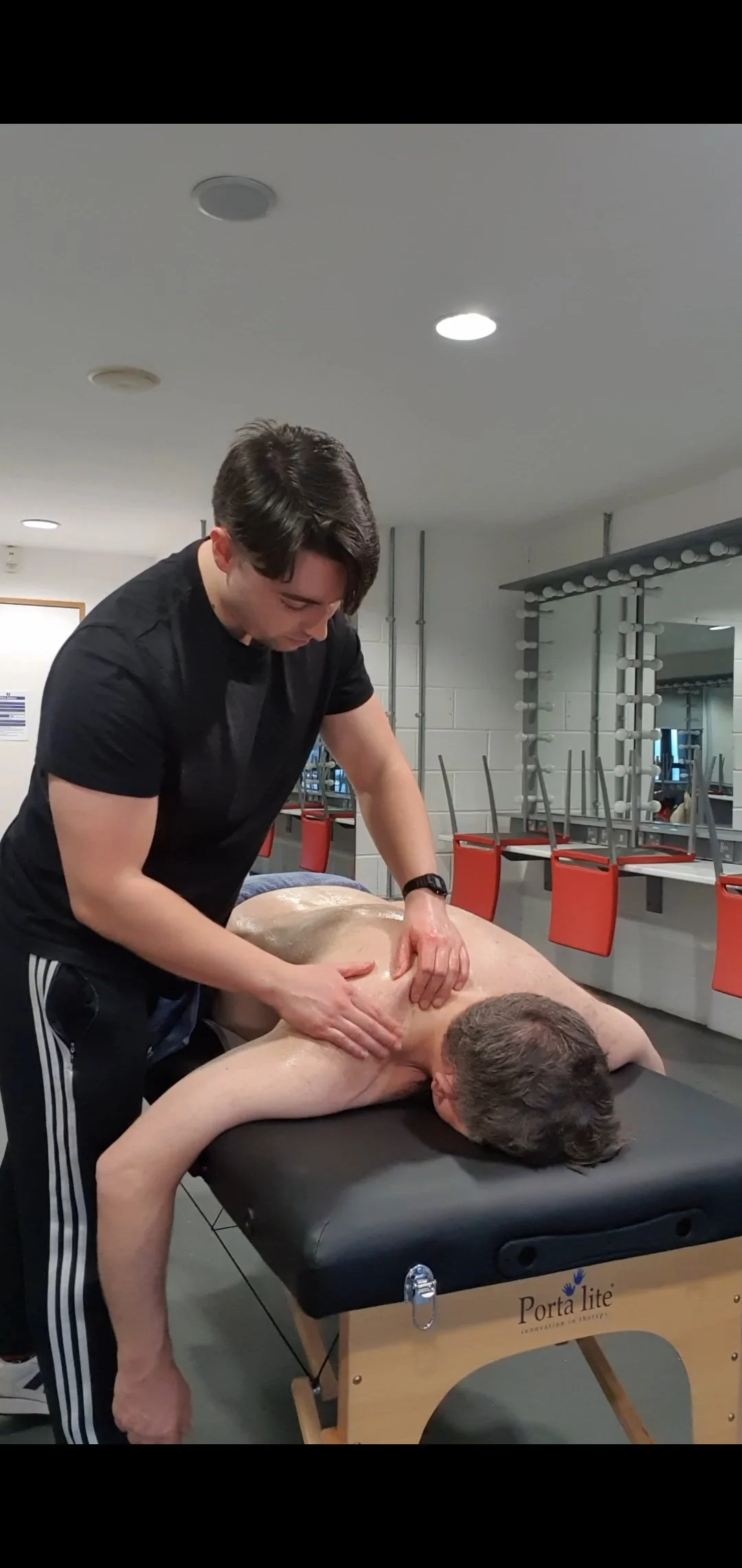 A massage therapist providing a back massage to a man lying face down on a massage table in a fitness or therapy room.