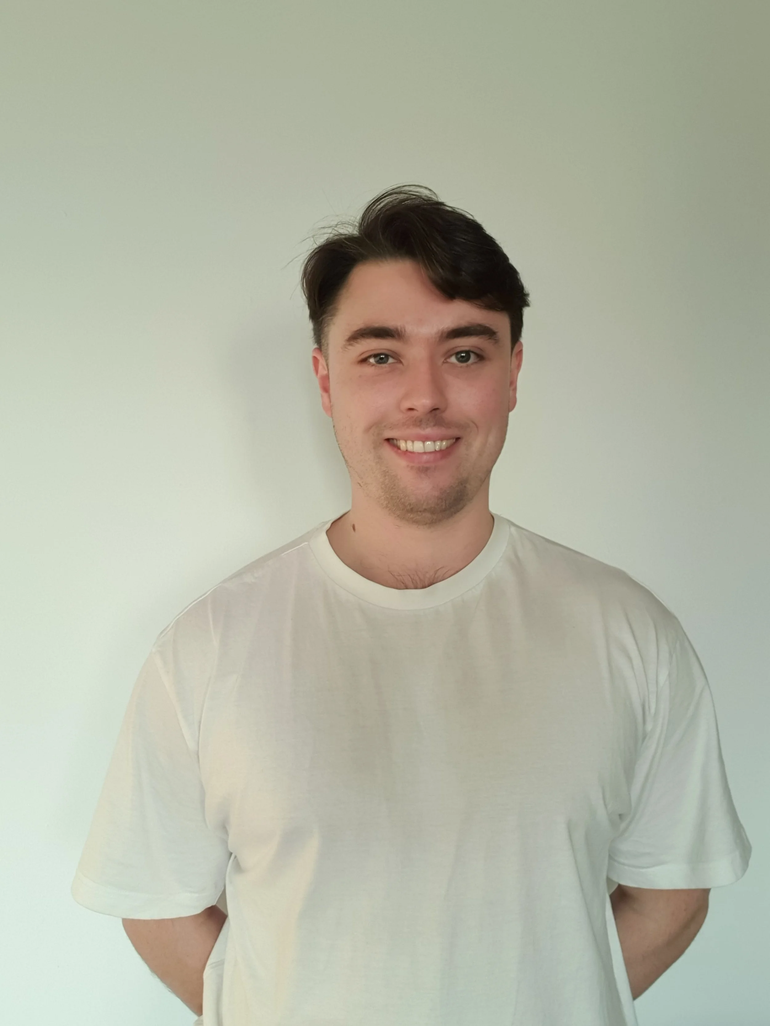 A smiling young man with dark hair wearing a plain white t-shirt standing against a light-colored wall.