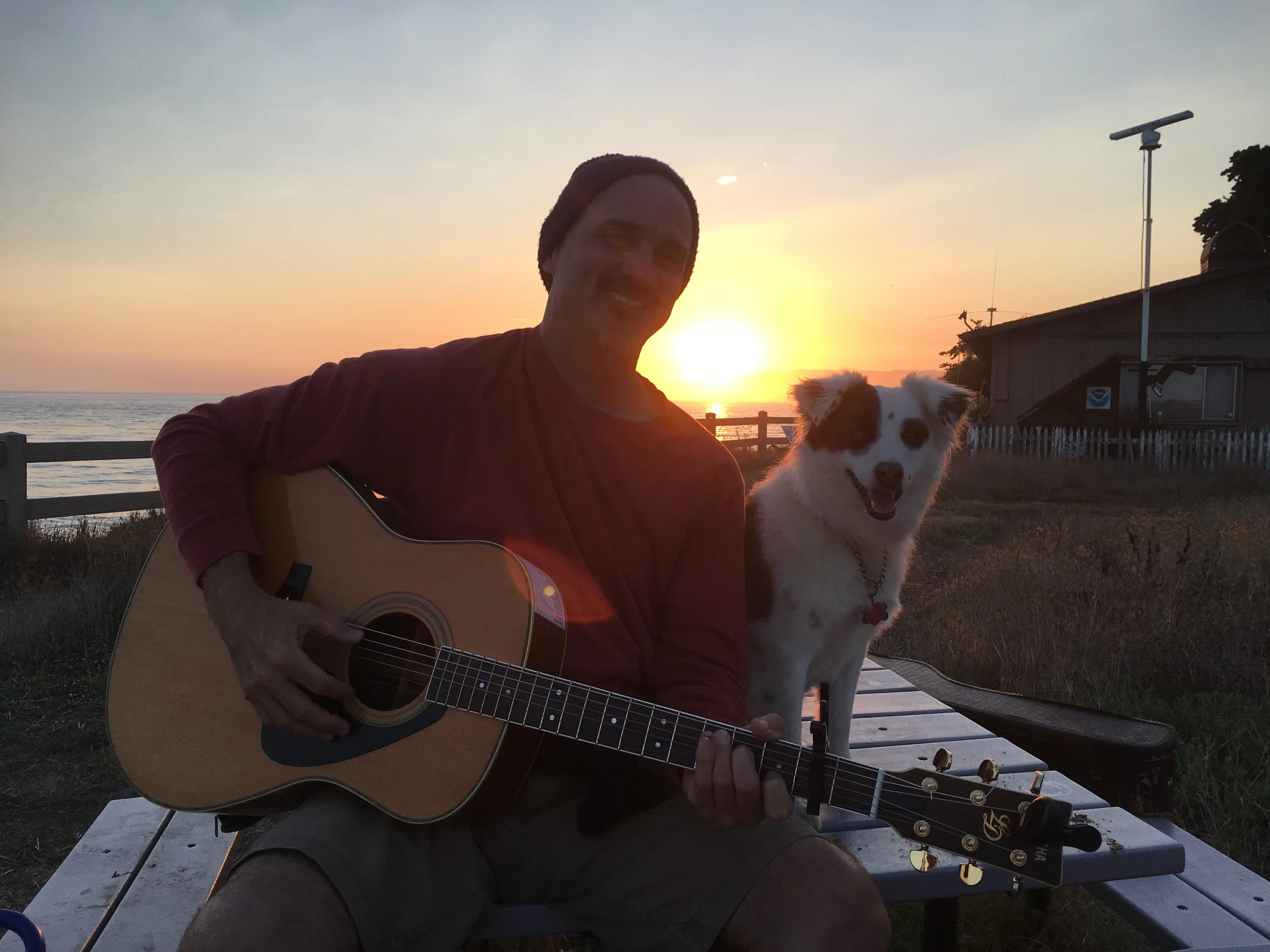 Eric Zobel seated with a guitar next to his dog, with the sun setting over the water in the background