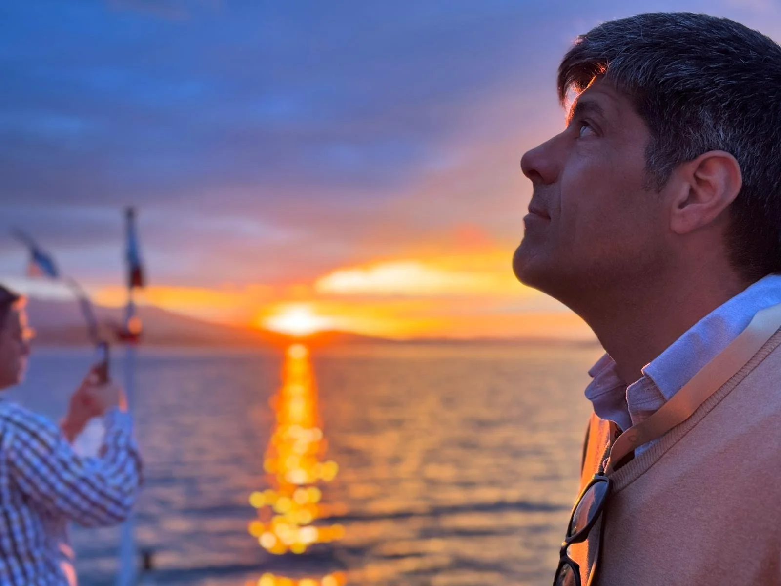 Homem olhando para o horizonte durante o pôr do sol no mar, ao lado de uma pessoa segurando uma vela.