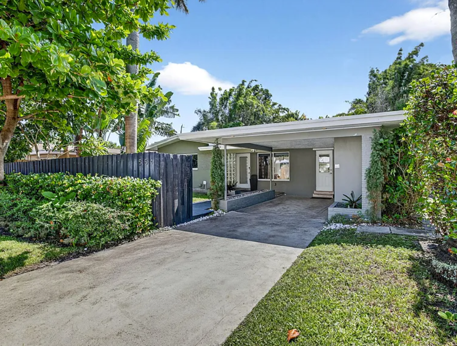 Modern single-story house with a concrete driveway, surrounded by green shrubs and trees, featuring a small porch area with covering, and a white door, under a blue sky.