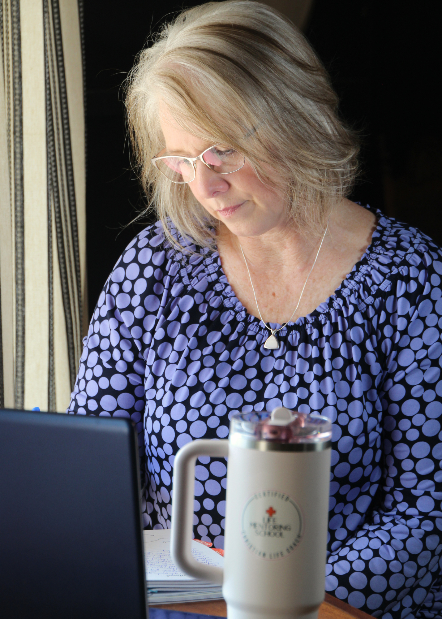 A woman with blonde hair, glasses, and a blue polka dot blouse sitting at a desk, working or reading in front of a laptop with a notebook and a white insulated mug.
