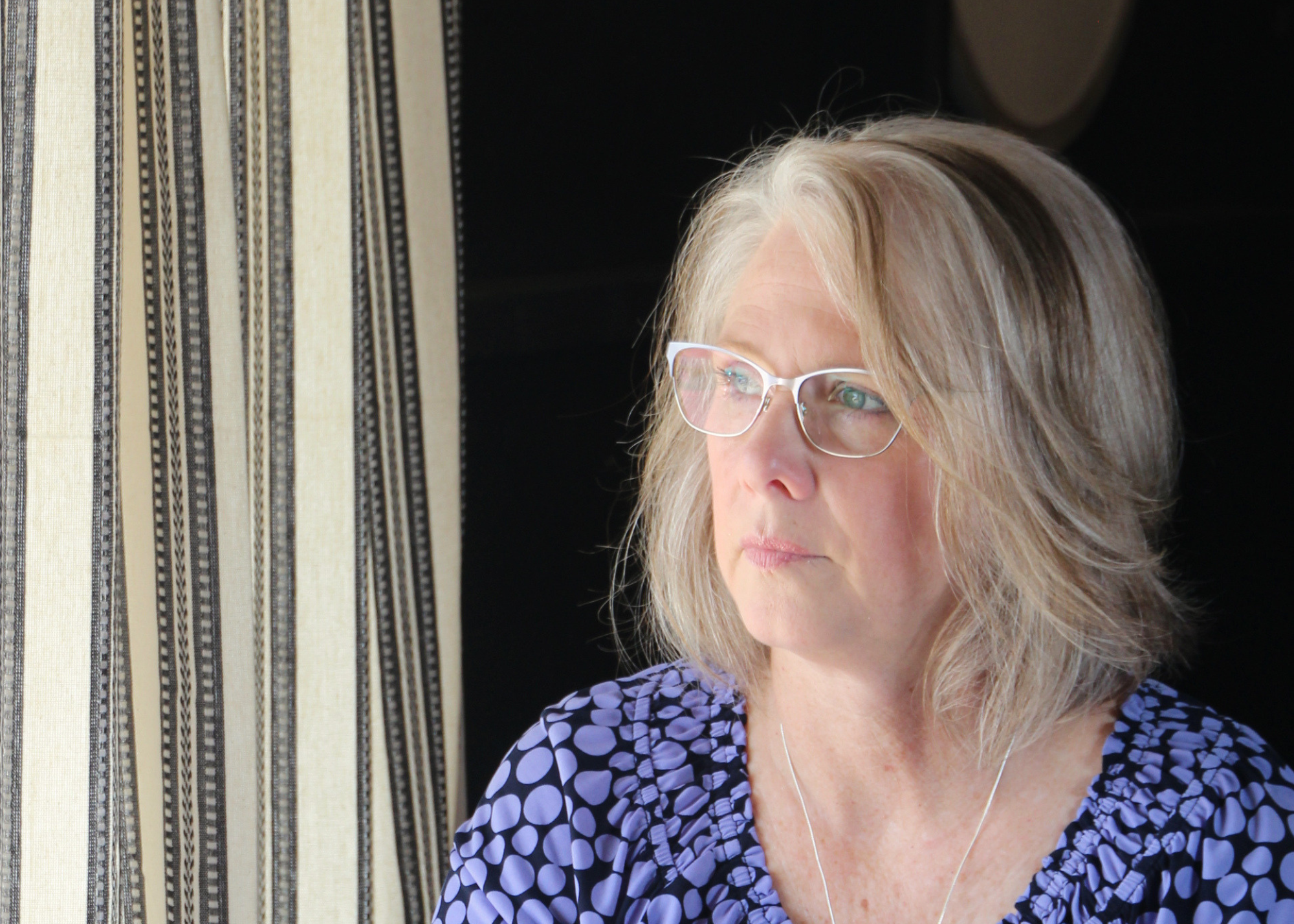 A woman with blonde hair and glasses looking to her right, wearing a polka dot blouse, sitting near a window with striped curtains.